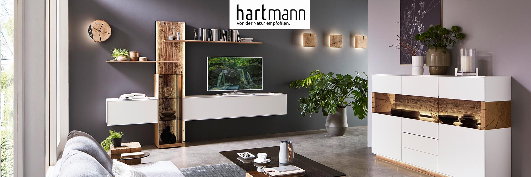 Hartmann BENJO - Wohnzimmermöbel aus massiver Birke weiß lackiert kombiniert mit Kerneiche Umato. Ambientebild zeigt beleuchtetes Holzpaneel eingefasst von zwei, weißen Hängelowboards, rechts ein Highboard mit Glastüren.