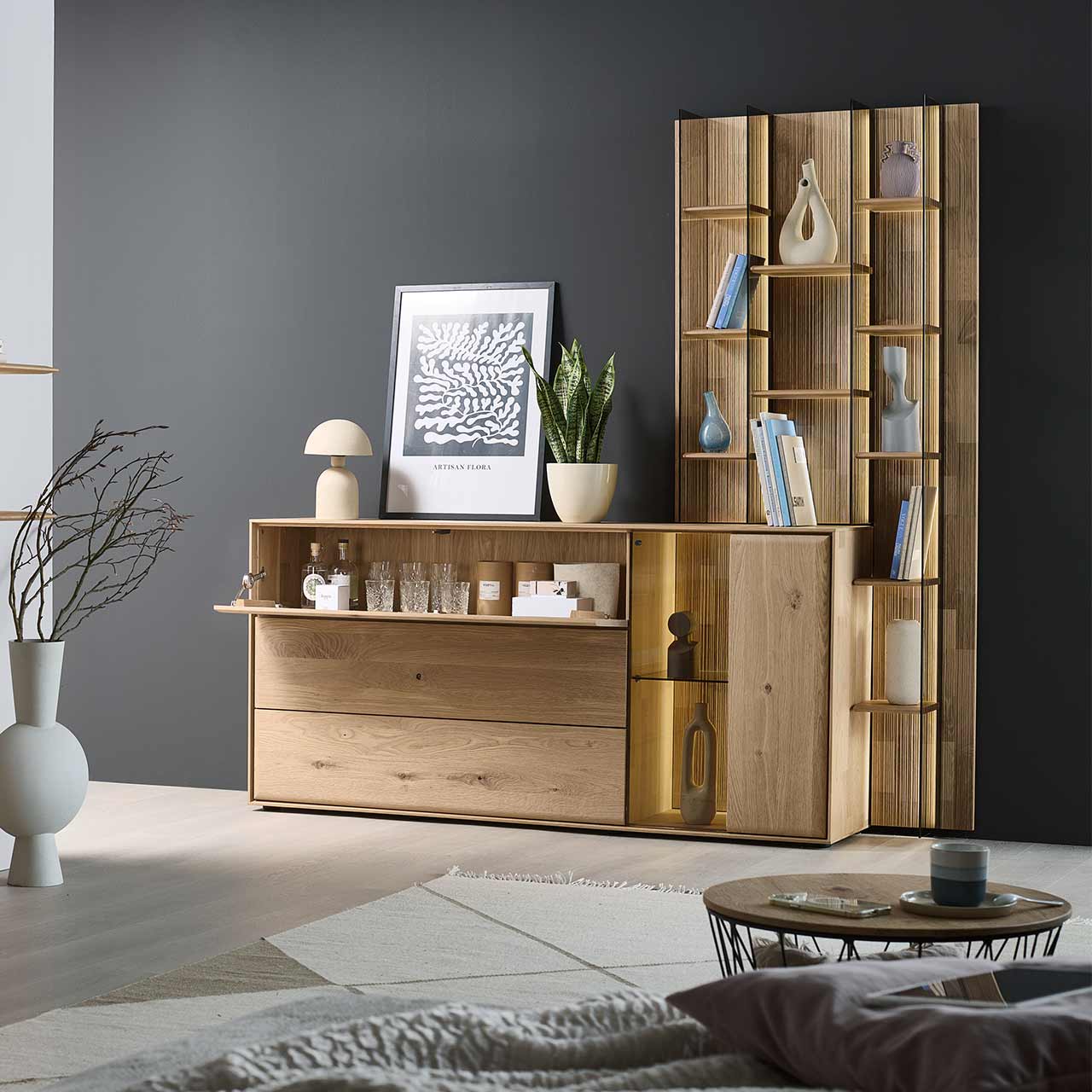 Modernes Sideboard aus massiver Kerneiche Natur mit beleuchteter Glas/Holztür, offener Holzklappe oben und zwei Holzschubkästen. Das Sideboard ist integriert in ein rechts stehendes, hohes Wandregal mit Holzböden und Glasseiten.