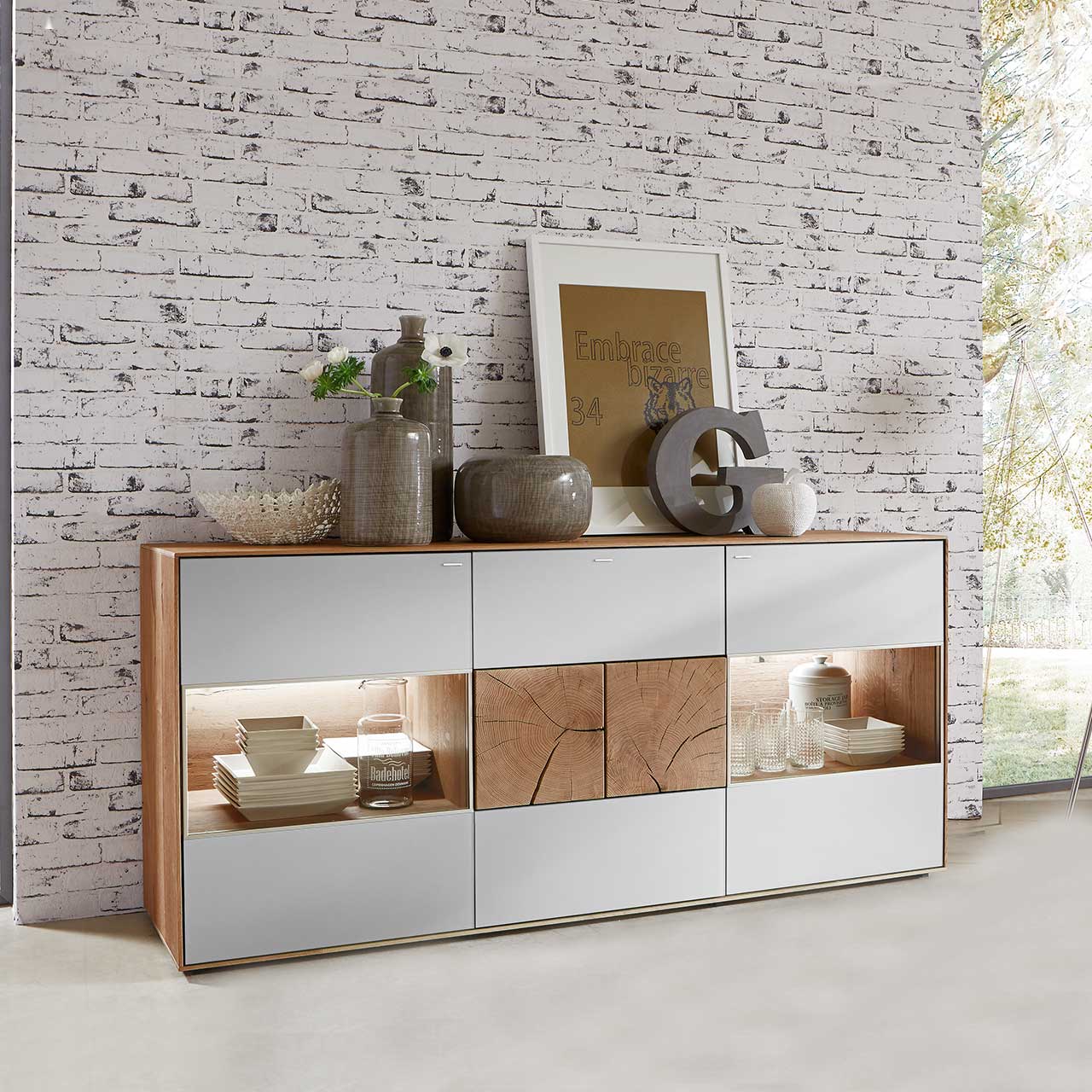 Modernes Sideboard aus massiver Kerneiche Umato mit weißfarbenen Mattglasfronten. Rechts und links mit beleuchtetem Glasausschnitt, mittig mit zwei dekorativen, rechteckigen Hirnholzscheiben - vor hellgrauer Steinwand-Tapete.