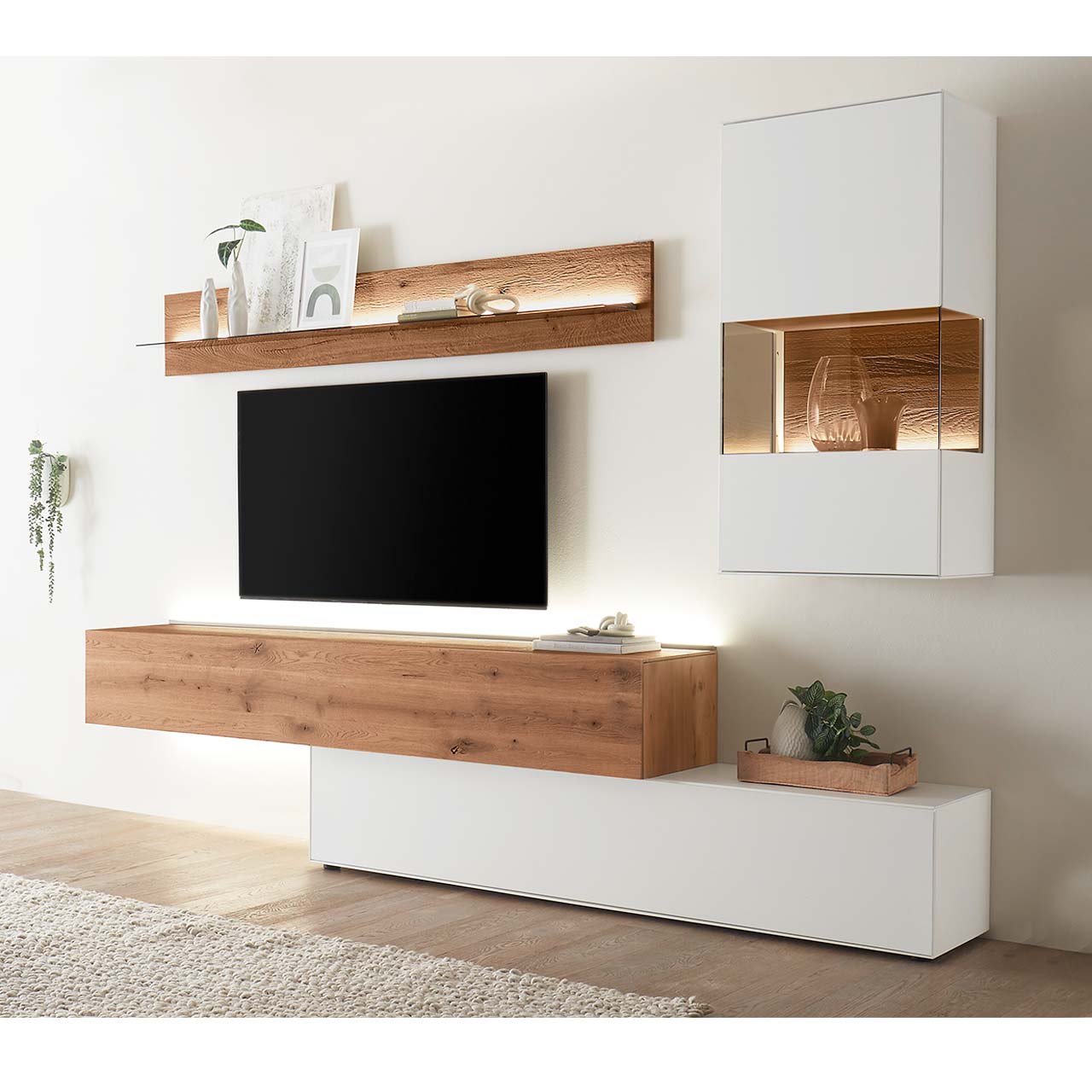 Moderne Wohnwand aus weiß lackierter Birke und dunkler Kerneiche Umato bestehend aus einem Hängelowboard seitlich eingefasst in einen weißen, langen Unterschrank, daneben eine weiße Hängevitrine neben einem Wandregal mit Glasablageboden. 