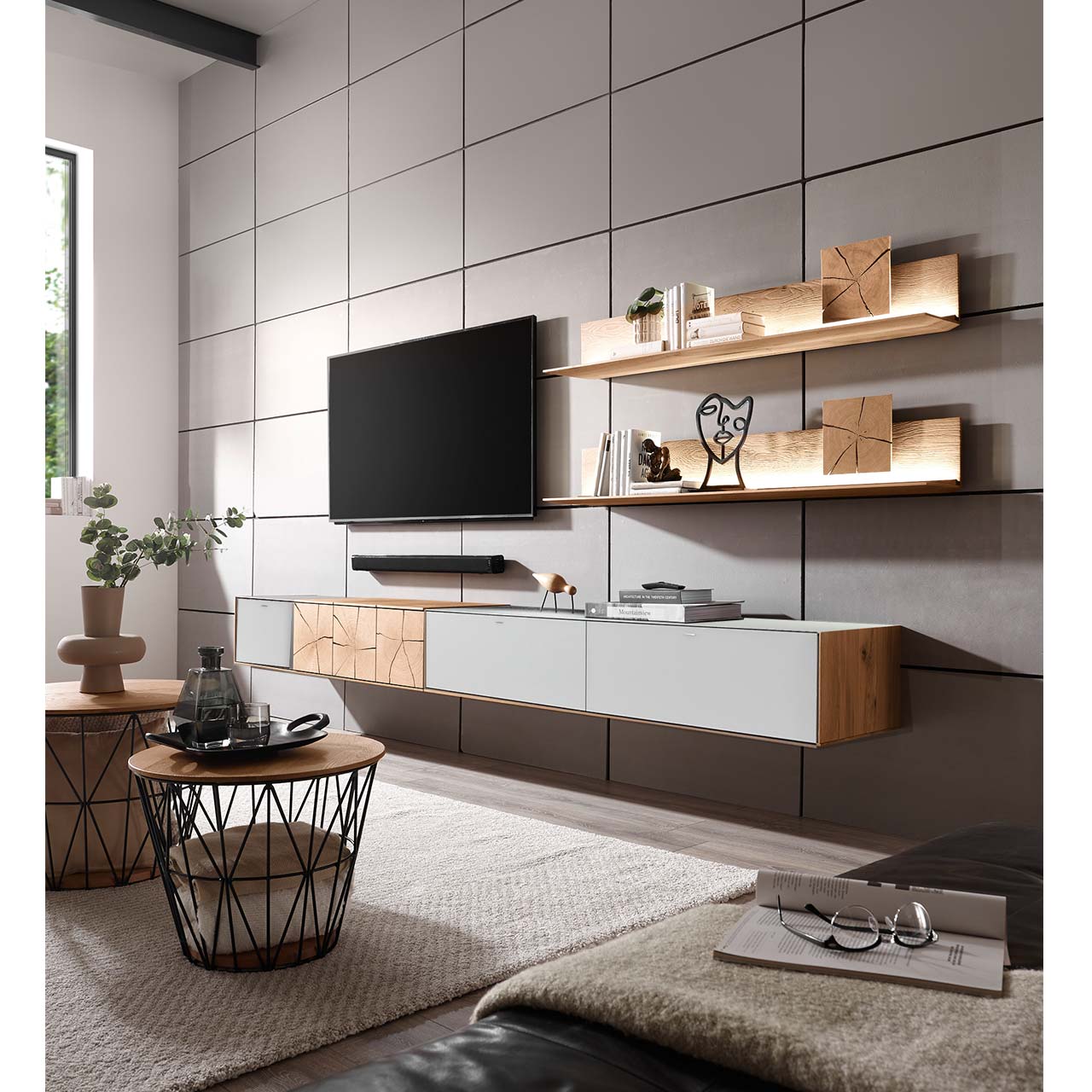 Wohnzimmer mit TV und moderner Wohnwand aus massiver Kerneiche Umato mit Mattglas weiß Fronten, mit langen Hängelowboard und zwei Wandpaneelen mit Hirnholzscheiben - an gefliester grauer Wand. Im Vordergrund zwei runde Couchtische mit Korbgestell.