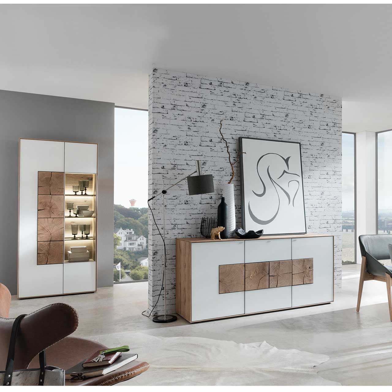 Milieubild Hartmann CAYA - Standvitrine 7170-0096W li. Flur mit modernem Sideboard aus massivem Holz mit Mattglas weiß Front und vier Hirnholzscheiben. Im Hintergrund an einer Wand stehend eine Strandvitrine in Mattglas weiß und beleuchtetem Glasausschnitt mit drei Glasböden.
