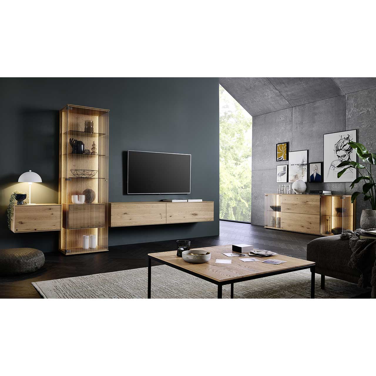 Eingerichtetes Wohnzimmer mit dreiteiliger Wohnwand aus Kerneiche Natur: zwei Hängelowboards und eine Standvitrine vor dunkelgrauer Wand. Rechts beleuchtetes Sideboard mit Glastüren, im Vordergrund rechteckiger Couchtisch.