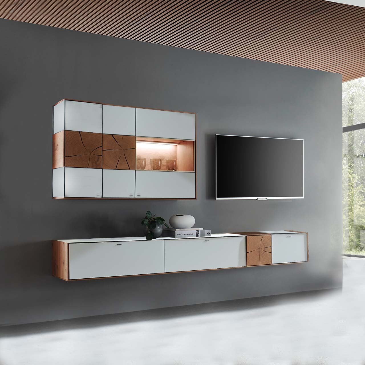 Massivholz Wohnwand aus Kerneiche mit Mattglas weißen Fronten. Unten ein langes, schmales Hängelowboard mit dekorativen Hirnholzscheiben, darüber die passende, beleuchtete Hängevitrine. Rechts daneben hängender Fernseher - vor dunkelgrauer Wand.