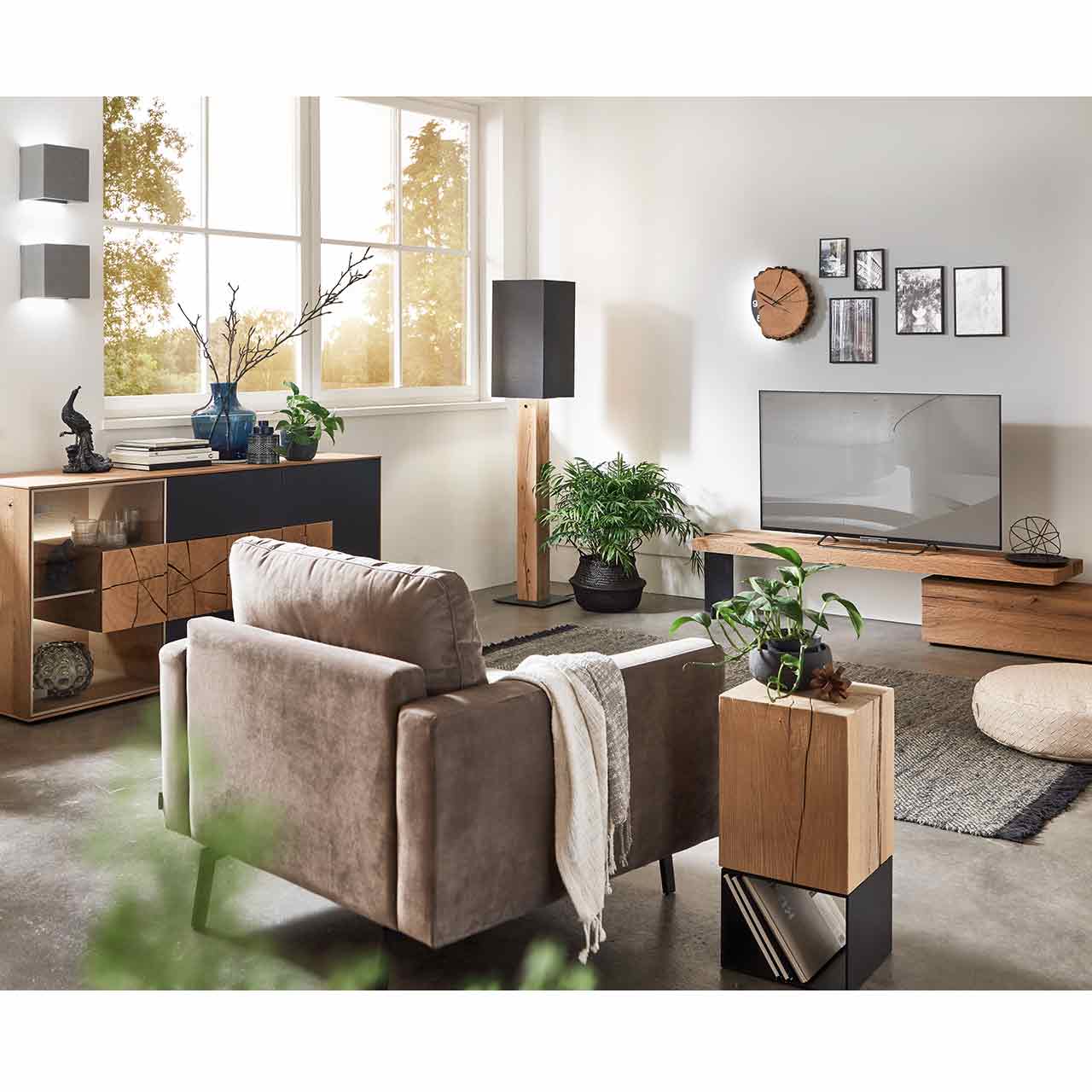 Milieubild Wohnzimmer - Hartmann NATURSTÜCKE 7610-1066 und 1016 Eingerichtetes Wohnzimmer mit modernem Sideboard aus Kerneiche, Stehlampe mit Stofflampenschirm und Holzfuß und einer Holzwanduhr. Im Vordergrund ein passender Beistelltisch mit Holzkorpus und Metallgestell.