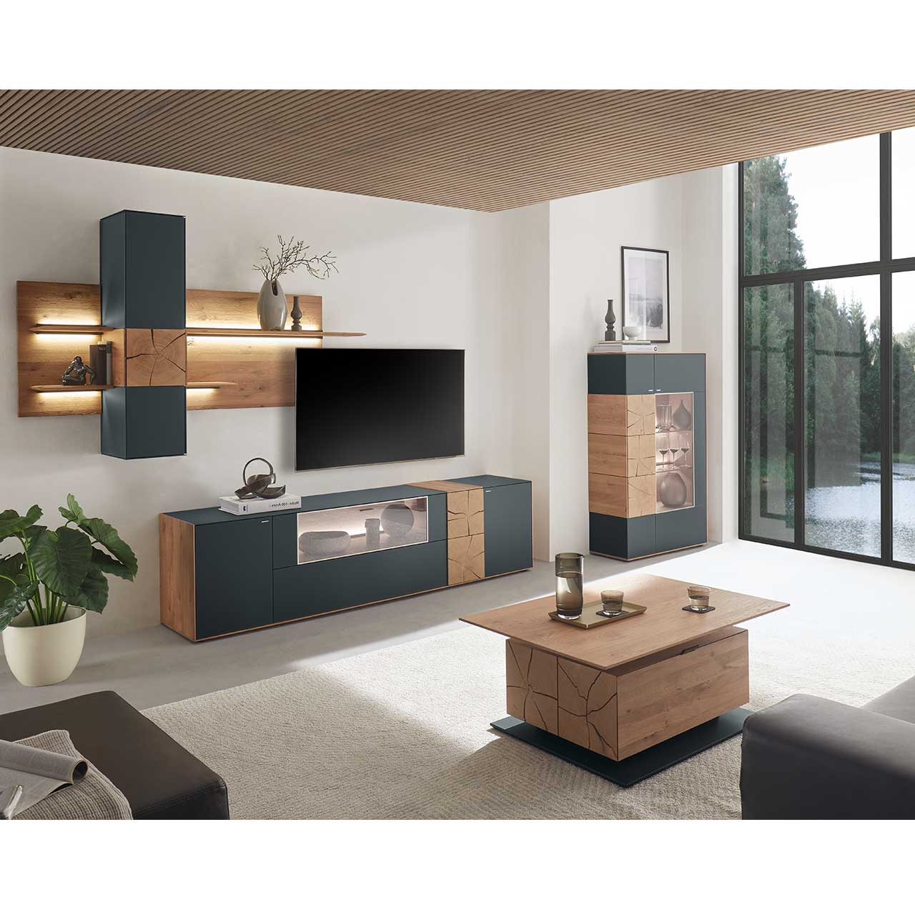 Wohnzimmer mit moderner Massivholz Wohnwand mit Mattglas anthrazit Fronten, mit Lowboard mit Glasausschnitt sowie Wandpaneel mit Regalböden und Hängeschrank. Rechts passendes Highboard mit Glasfront und Hirnholzscheiben. Im Vordergrund Holz-Couchtisch. 