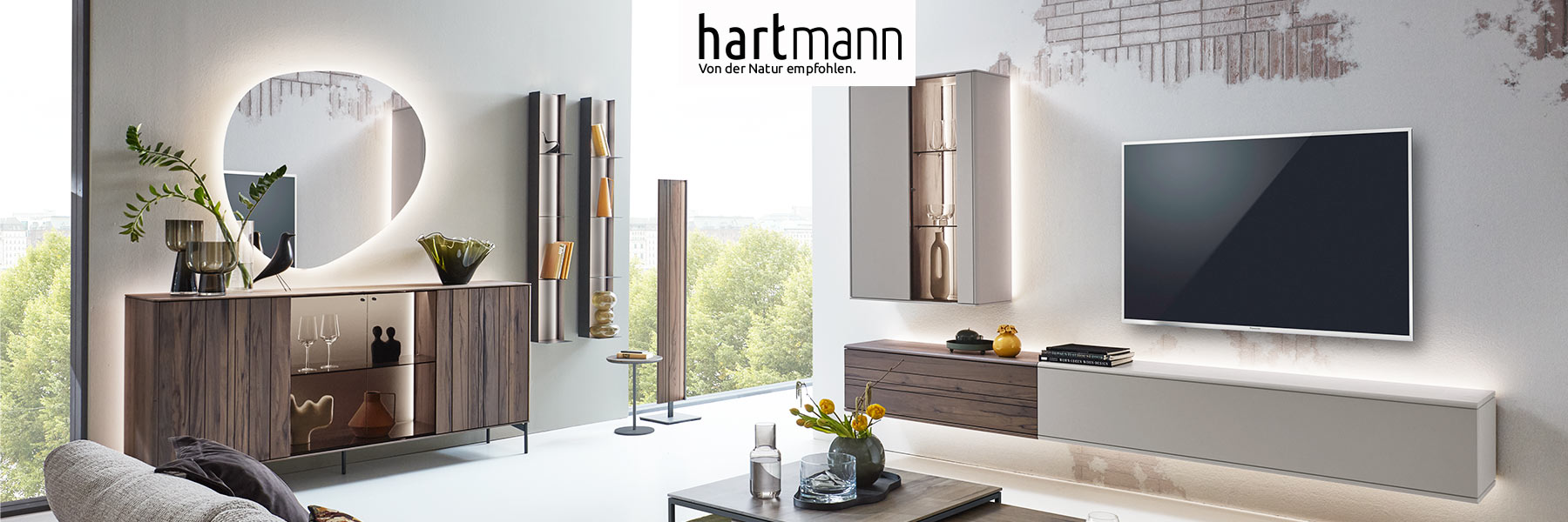 Hartmann AUDRA - Moderne Möbel aus massiver Altholzbuche Uyaa kombiniert mit sandfarbenen Lackfronten. Ambientebild zeigt Wohnzimmer mit dreiteiliger Wohnwand, Couchtisch, zwei Hängeregale und Sideboard mit Metallfüßen darüber moderner Spiegel.