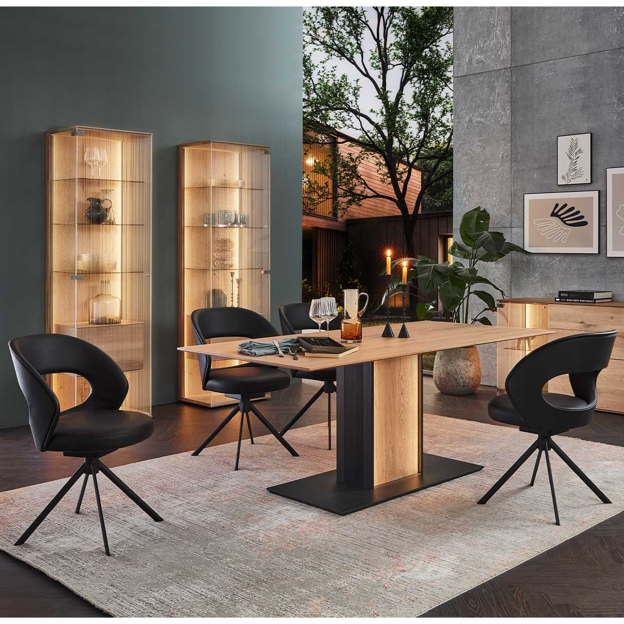 Eingerichtetes Esszimmer mit modernem Esstisch aus massiver Kerneiche Natur und vier Stuhlen im schwarzem Leder auf Metallgestell. Links stehen zwei beleuchtete Standvitrinen, rechts das passende Sideboard.