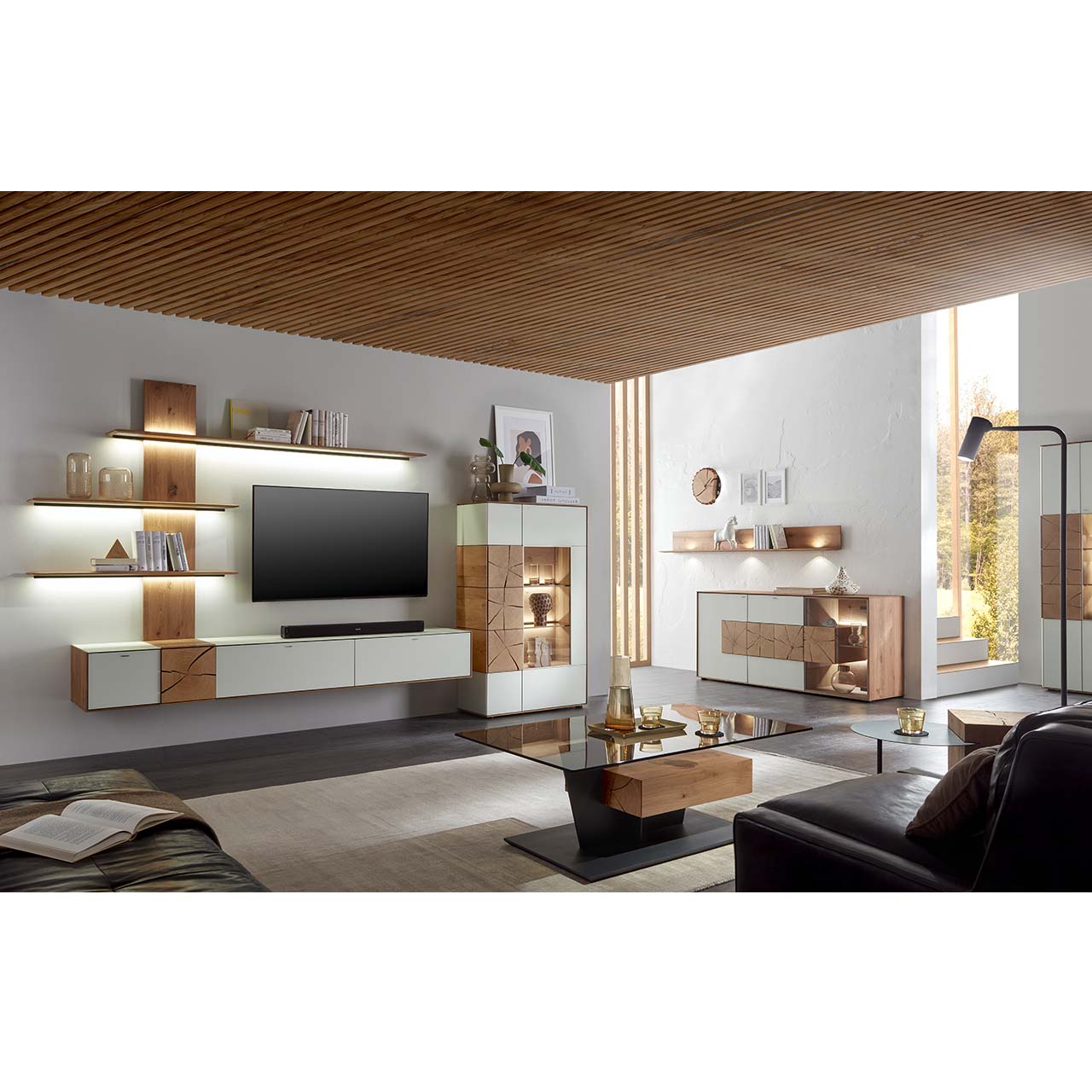 Wohnzimmer mit TV und moderner Wohnwand aus massiver Kerneiche Umato mit Mattglas weiß Fronten und Hirnholzscheiben, mit Hängelowboard, Wandpaneel mit drei Regalböden und Highboard mit Glasfront. Im Vordergrund Couchtisch mit rechteckiger Glasplatte.