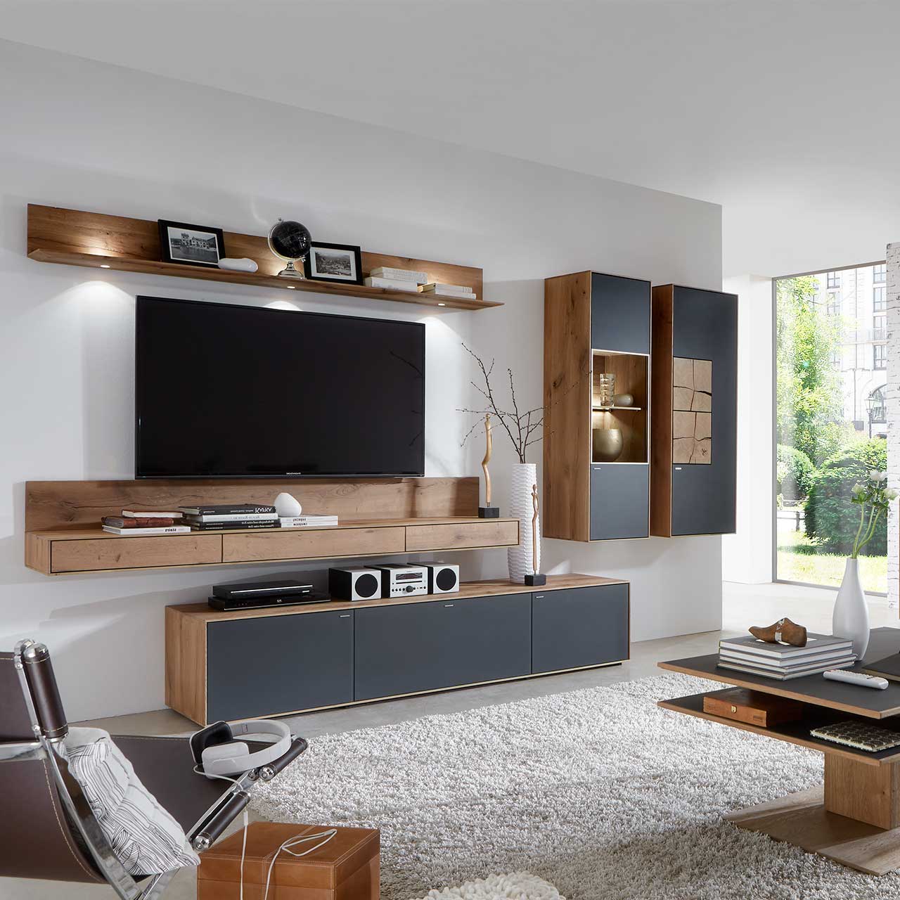 Wohnzimmer mit TV und moderner Wohnwand aus massiver Kerneiche Umato mit Mattglas anthrazit Fronten: Beleuchtetes Wandregal, langes Lowboard, beleuchtete Hängevitrine und Hängeschrank mit dekorativen Hirnholzscheiben.