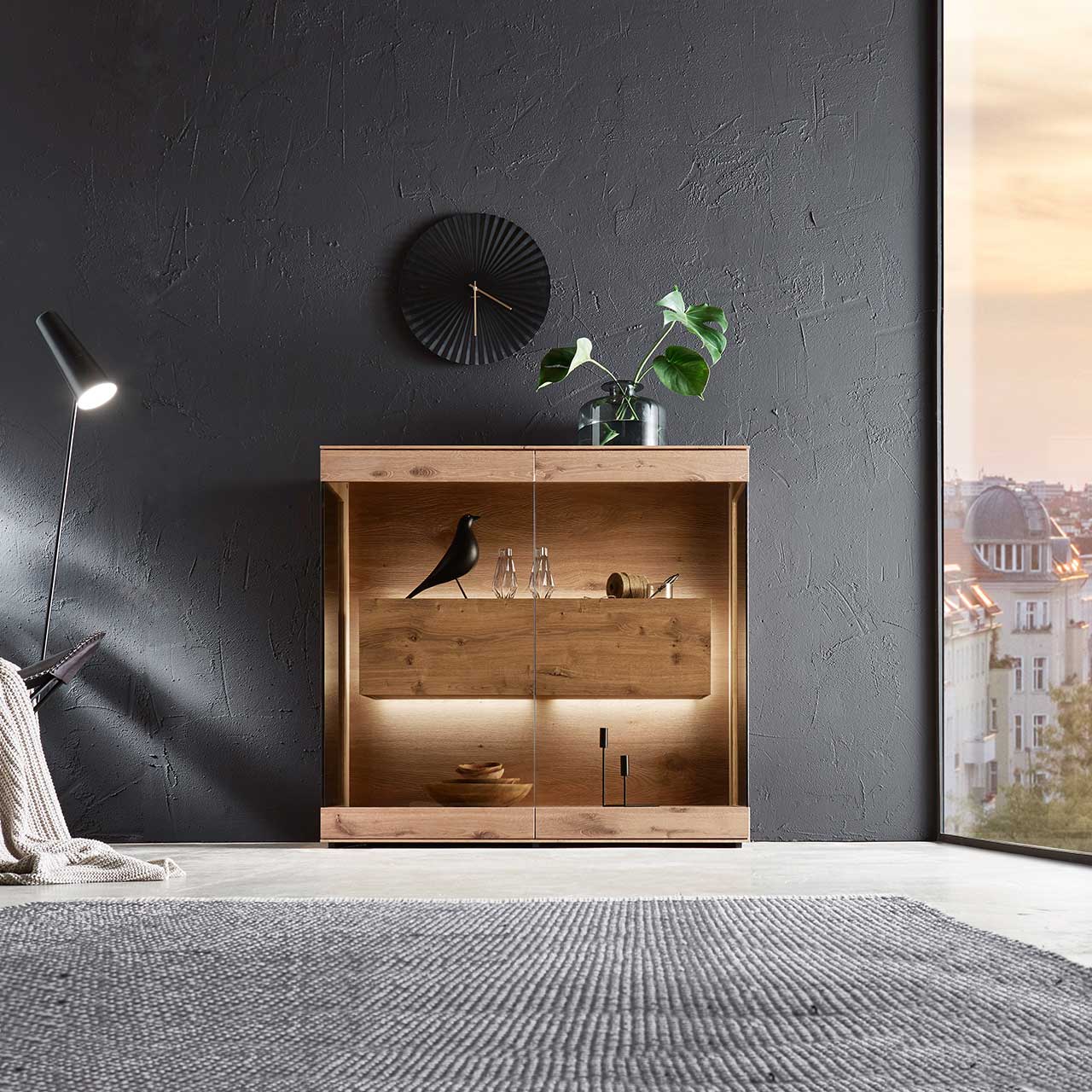Edles Highboard aus massiver Kerneiche Umato mit großen über Ecke verlaufenden Glastüren in Parsolbronze. Innenliegend ein großer, beleuchteter Hirnholzblock mit Doppelschubkasten - an dunkelgrauer Wand stehend.