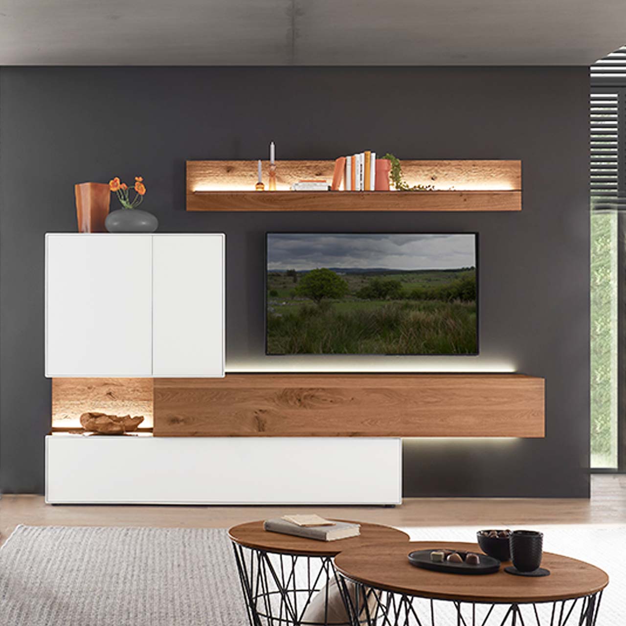 Moderne Wohnwand aus massiver Birke weiß lackiert mit Kerneiche Umato bestehend aus einem Holz-Hängelowboard seitlich eingefasst in einen weißen Unter,- und Hängeschrank mit beleuchteter Rückwand und einem Holz-Wandpaneel mit Glasablageboden.