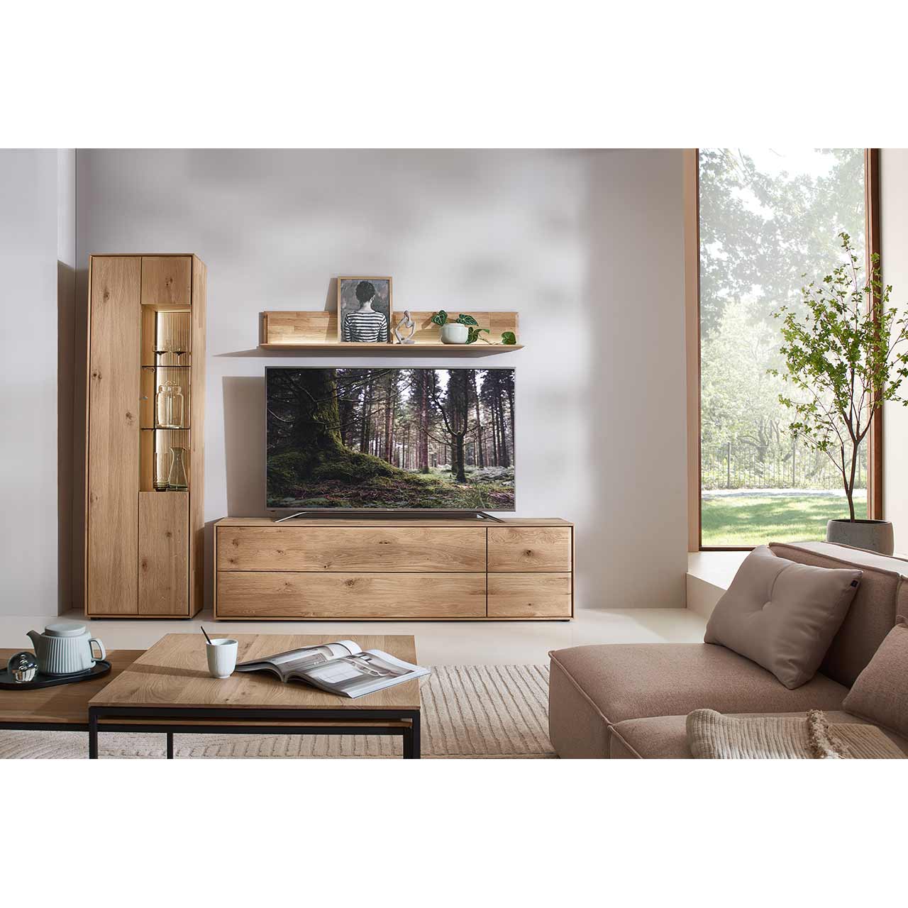 Modernes Wohnzimmer mit dreiteiliger Massivholzwohnwand bestehend aus einer beleuchteten Standvitrine, einem breitem Wandregal mit Holzboden und geschroppter Rückwand und einem Lowboard. Davor rechteckiger Holz-Couchtisch.