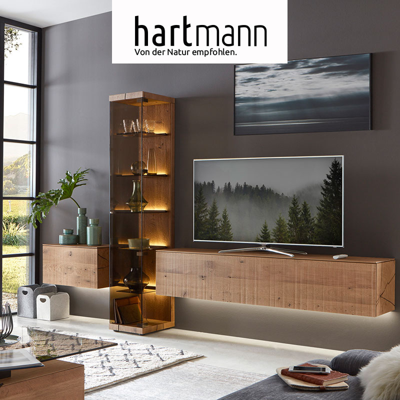 Hartmann VARA - Wohnzimmermöbel aus massiver Kerneiche Umato sägerau gebürstet kombiniert mit Metall Anthrazit Fronten. Ambientebild zeigt zwei schlichte Hängelowboards, dazwischen eine edle, beleuchtete Standvitrine aus Parsolglas - vor dunkler Wand