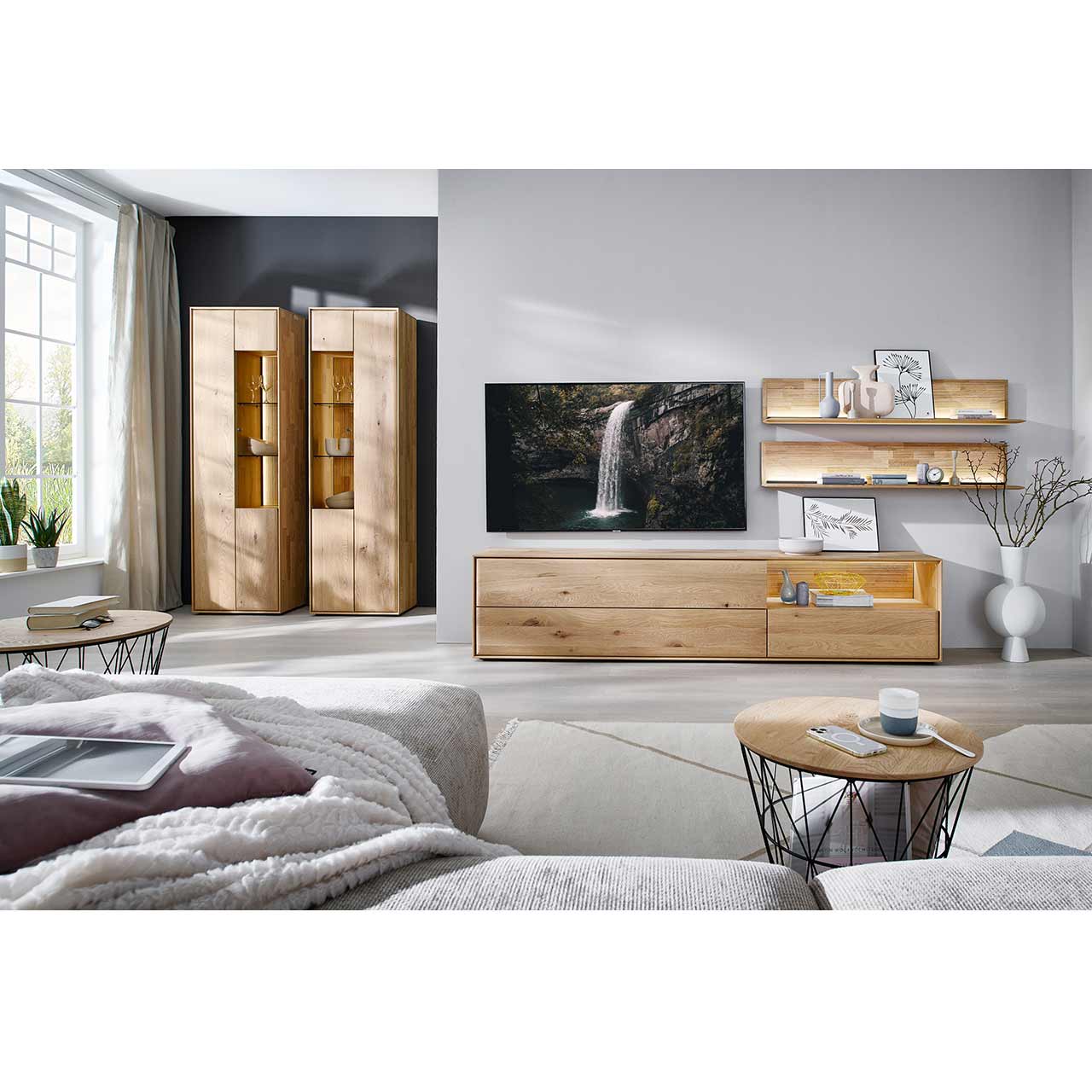 Wohnzimmer mit moderner Massivholzwohnwand bestehend aus langem Lowboard mit beleuchteter Nische und zwei beleuchteten Wandpaneelen mit Holzboden und geschroppter Rückwand. Davor runder Couchtisch, hinten links zwei passende Standvitrinen.