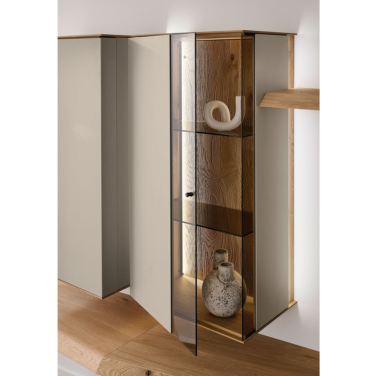 Detailansicht einer Vitrine aus massiver Kerneiche Natur mit Lackfronten in Sand. Mit geöffneter Glas-Lacktür, dahinter sichtbar die beleuchtete Riffholzrückwand sowie zwei Glasböden in Parsol Bronze.