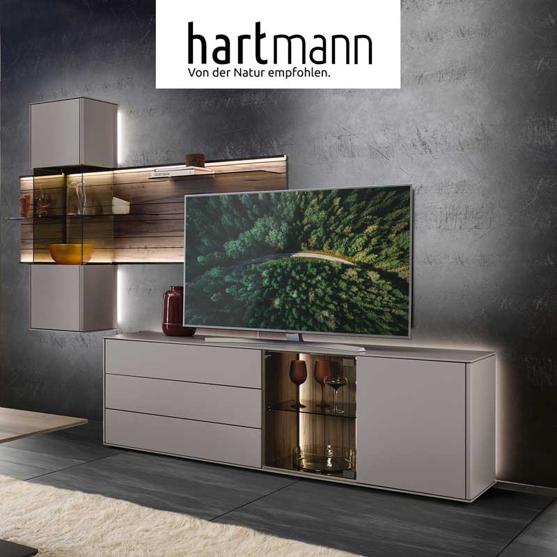 Hartmann Möbel - Möbelserie AUDRA Hartmann AUDRA - Wohnzimmermöbel aus massiver Altholzbuche Uyaa kombiniert mit modernem Lack. Ambientebild zeigt Lowboard mit Glasausschnitt und beleuchtete Hängevitrine in Lack sand eingefasst in braunes Wandpaneel mit Holzmaserung und Glasböden.