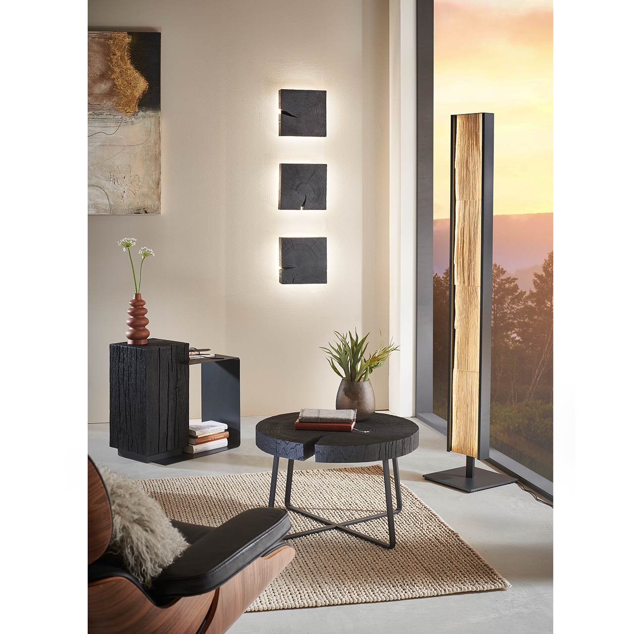 Milieubild Wohnzimmer - Hartmann NATURWERKE 9600C-0363 Runder Beistelltisch mit Tischplatte aus Kernesche anthrazit und Metallgestell, links daneben ein rechteckiger Beistelltisch mit dunklem Holzblock und Metall. Darüber hängend drei passende Wandleuchten, rechts eine Stehleuchte - im Wohnzimmer stehend.