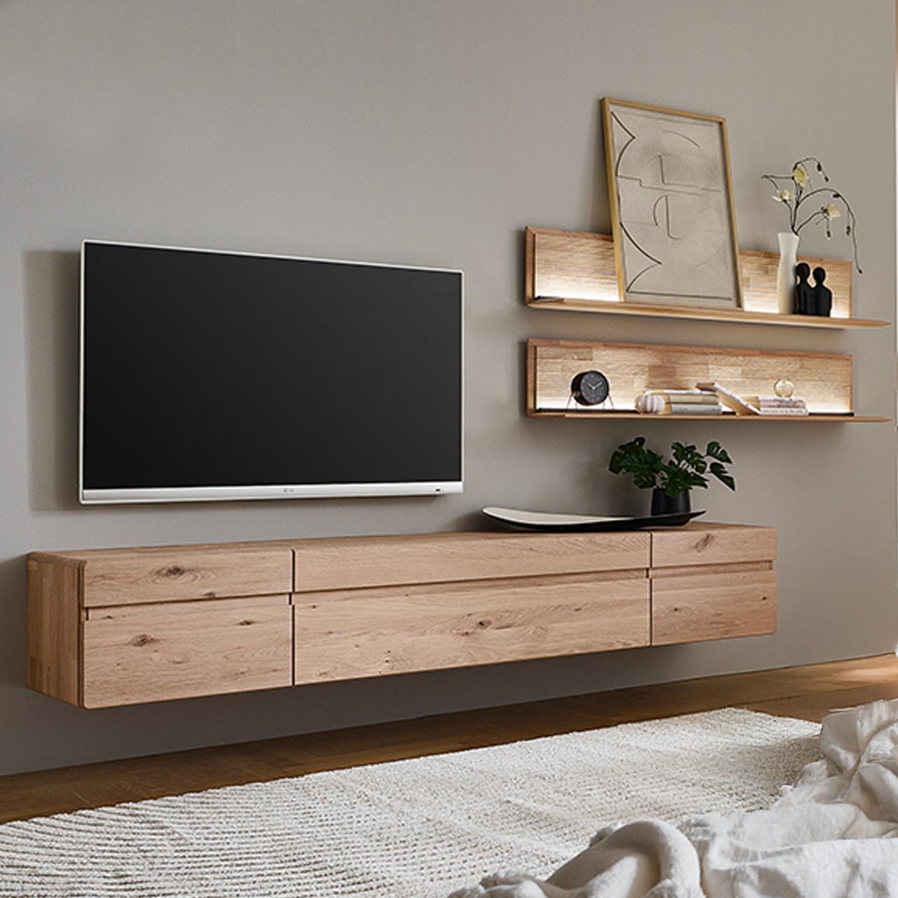 Moderne dreiteilige Wohnwand aus massiver Kerneiche Natur bestehend aus einem langen Hängelowboard mit Türen und Klappe sowie zwei übereinander hängenden Wandpaneelen mit Holzablageböden und beleuchteter, geschroppter Rückwand.