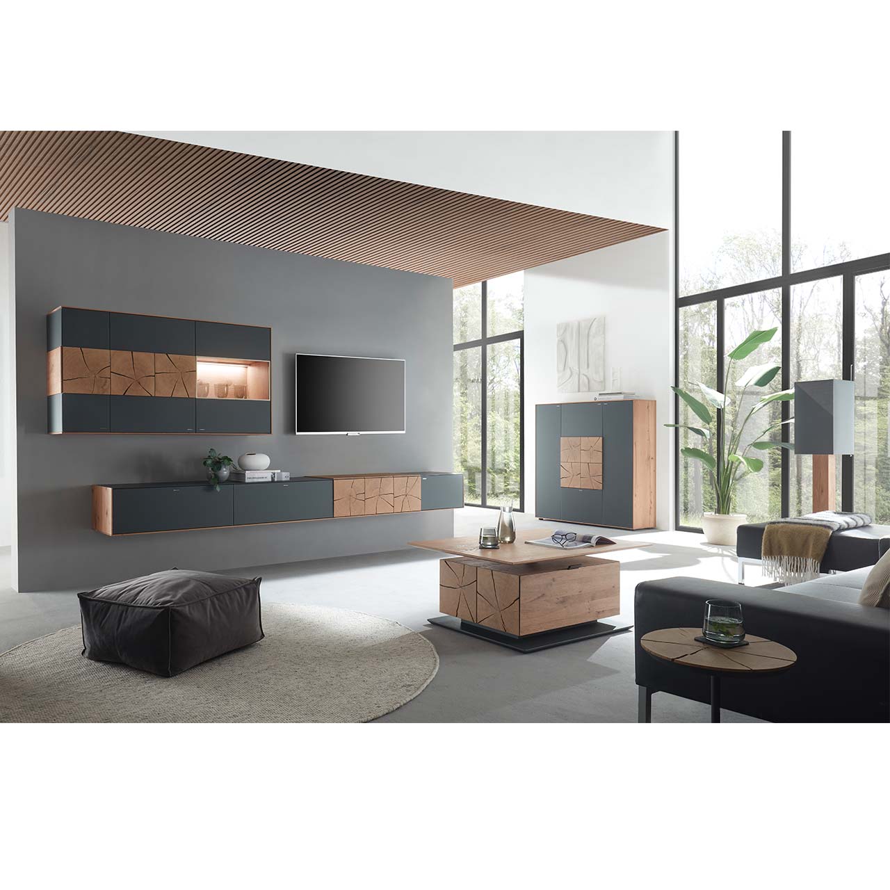 Wohnzimmer mit TV und moderner Wohnwand aus massiver Kerneiche und Mattglas anthrazit Fronten, mit schmalem Hängelowboard und beleuchteter Hängevitrine. Rechts das passende Highboard. Im Vordergrund ein rechteckiger Holz-Couchtisch.