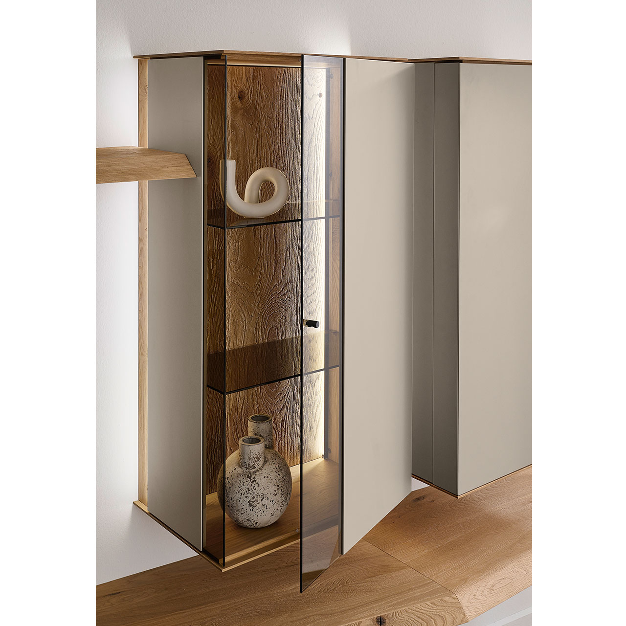 Hartmann Möbel SVEA - Hängevitrine 8660-6062 offen - Kerneiche Natur, Lack Sand Detailansicht einer Vitrine aus massiver Kerneiche Natur mit Lackfronten in Sand. Mit geöffneter Glas-Lacktür, dahinter sichtbar die beleuchtete Riffholzrückwand sowie zwei Glasböden in Parsol Bronze.