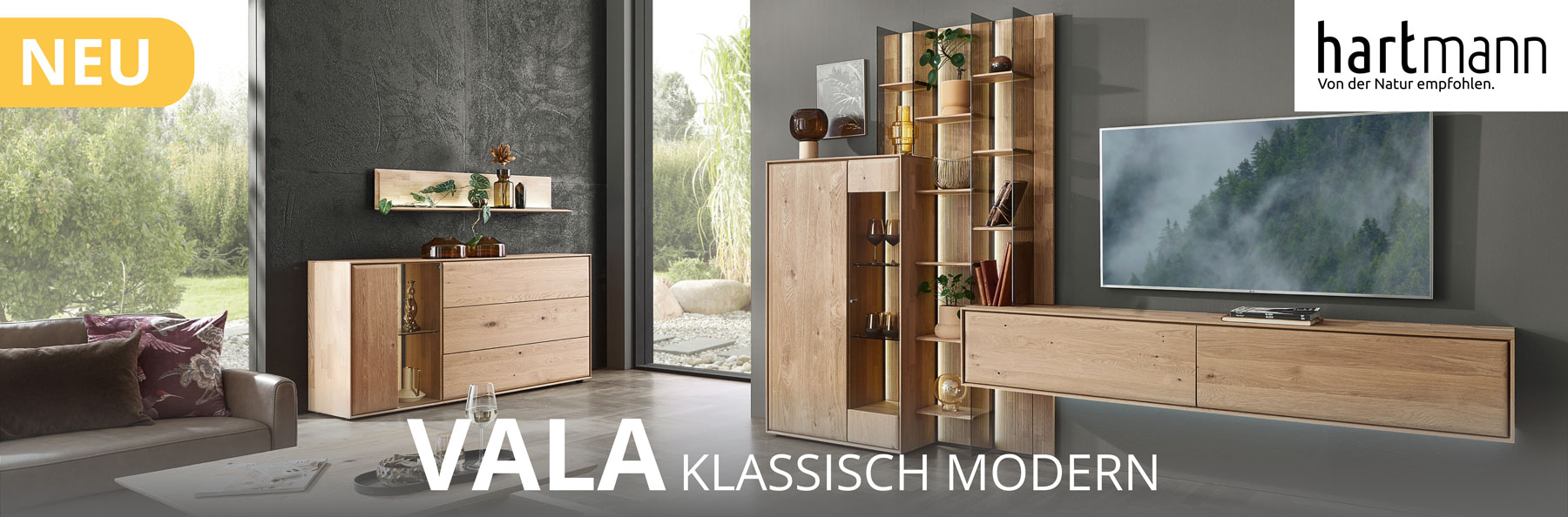 Hartmann VALA - Neue Möbelserie aus Kerneiche Natur kombiniert mit Parsolglas und geschoppten Rückwänden. Ambientebild zeigt schlichtes Lowboard, Wandpaneel mit beleuchteten Glasböden und integriertes Highboard. Weiter links Sideboard darüber Regal.