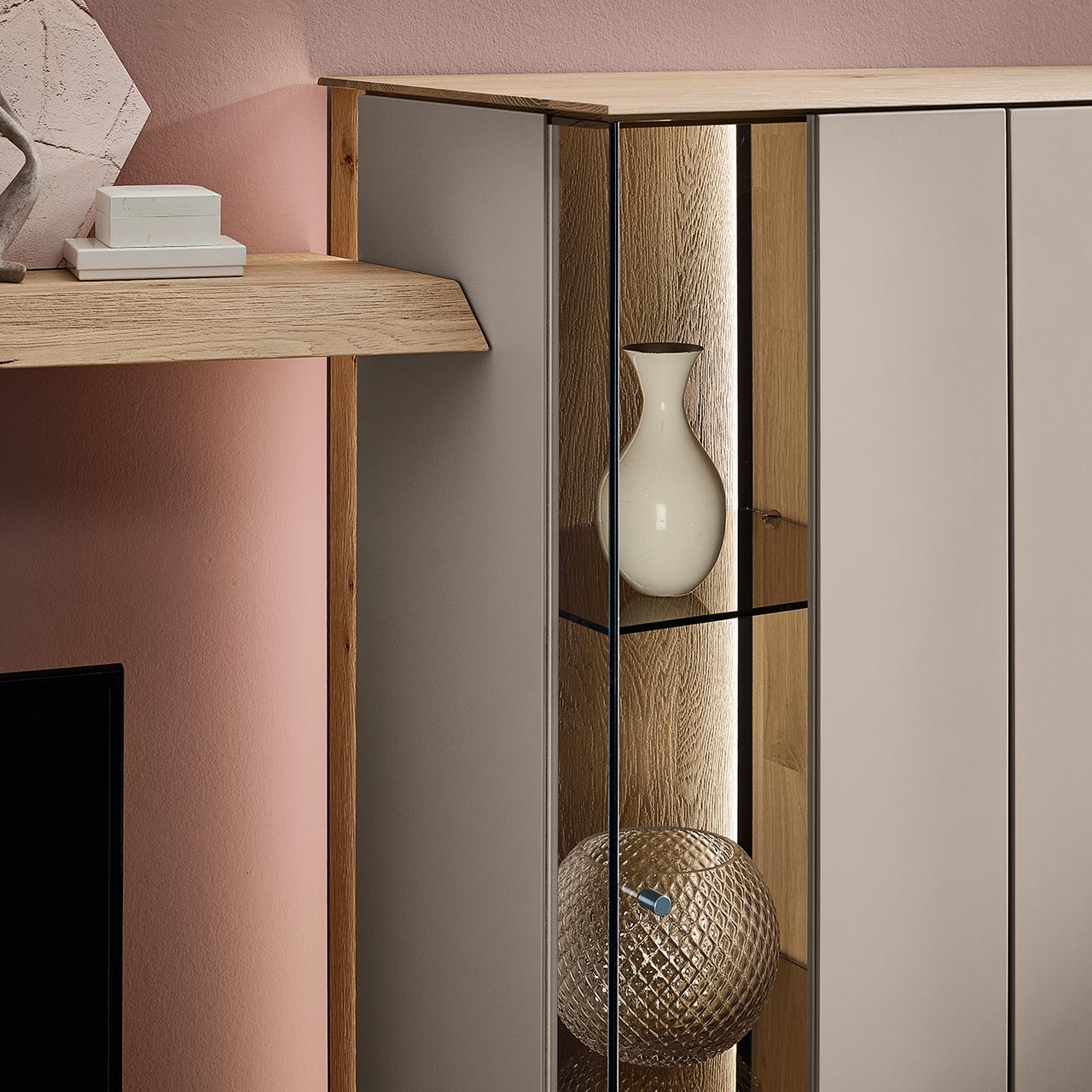 Detailansicht einer Vitrine aus massiver Kerneiche Natur mit Lackfronten in Sand mit Glas-Lacktür, dahinter sichtbar die beleuchtete Riffholzrückwand sowie zwei Glasböden in Parsol Bronze.