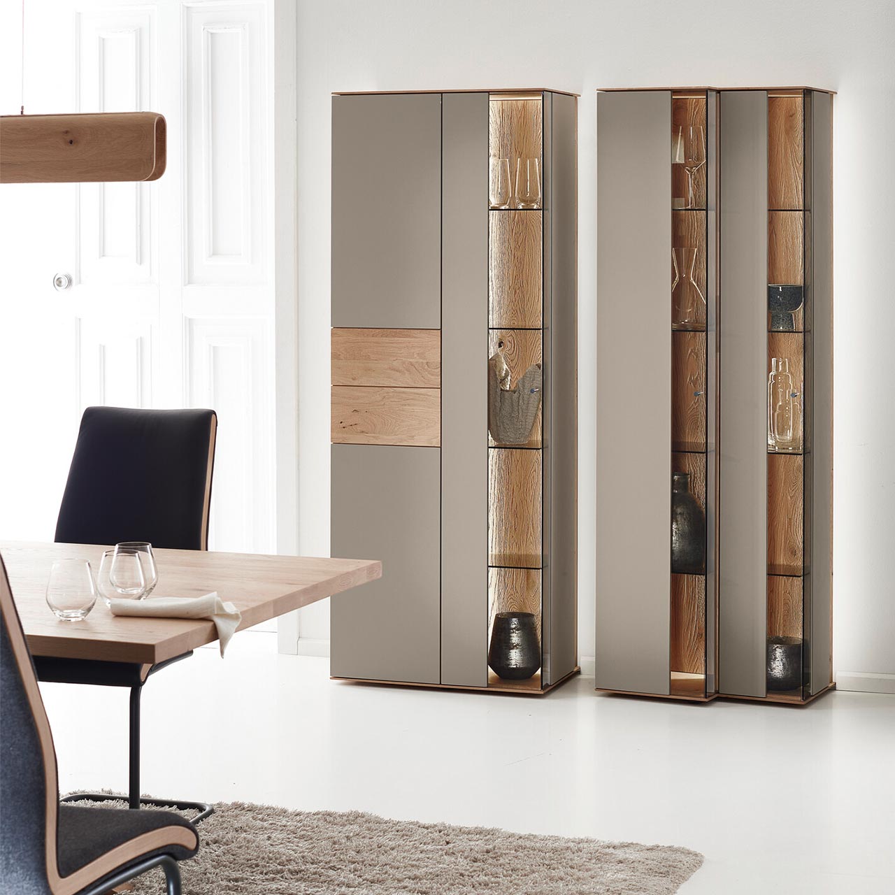 Hartmann Möbel SVEA - Standvitrinen 8660-0061 und 0071 - Kerneiche Natur, Lack Sand - Milieubild Moderne Standvitrinen aus massiver Kerneiche Natur mit Lackfronten in Sand. Mit edlen Glastüren in asymmetrischem Design und beleuchteten Riffholzrückwänden.