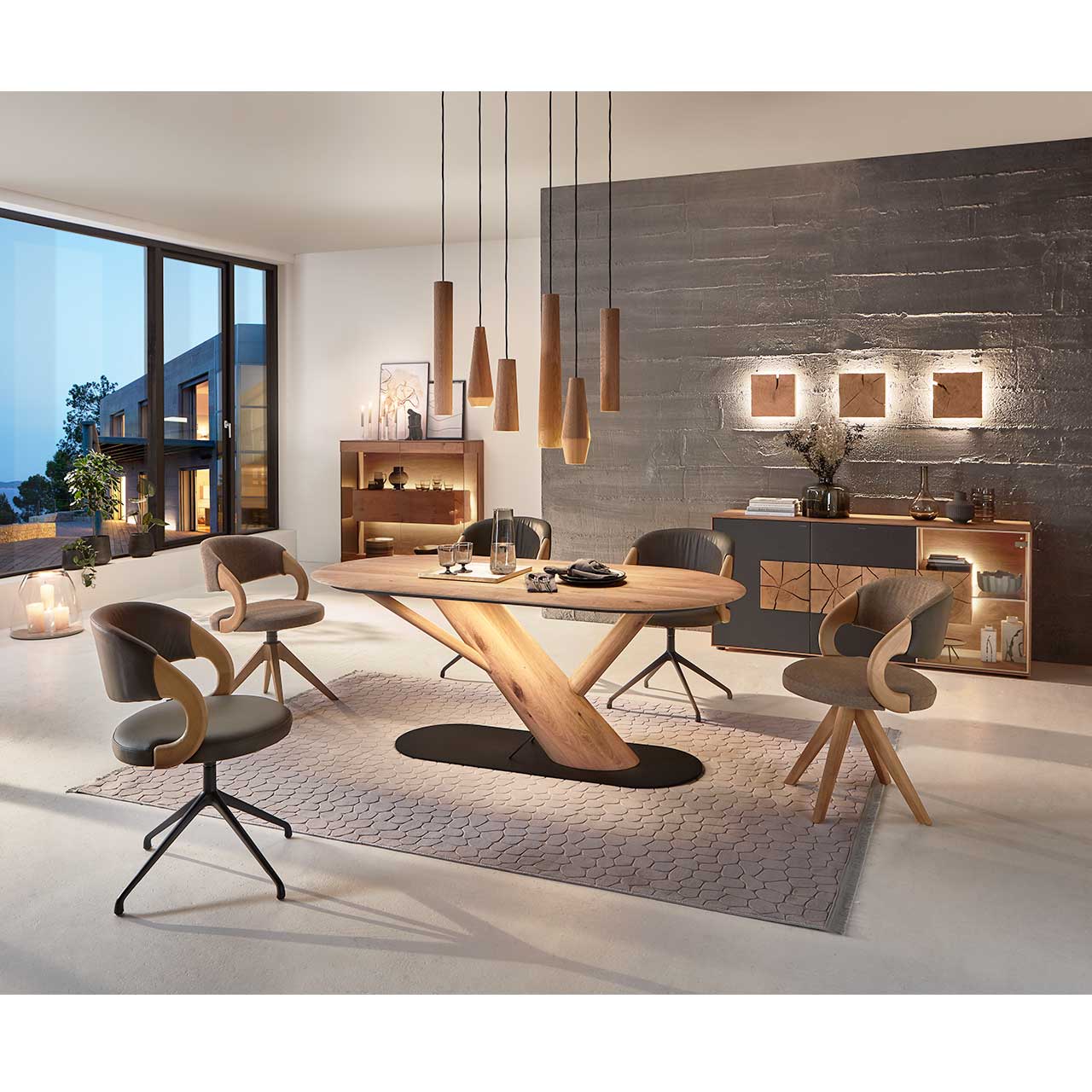 Modernes Esszimmer mit massivem, ovalem Holztisch, vier Stühlen auf Holz - und Metallgestell. Über dem Tisch hängen fünf Pendelleuchten mit Lampenschirmen aus Holz. Dahinter links an der Wand die passenden Wohnmöbel.