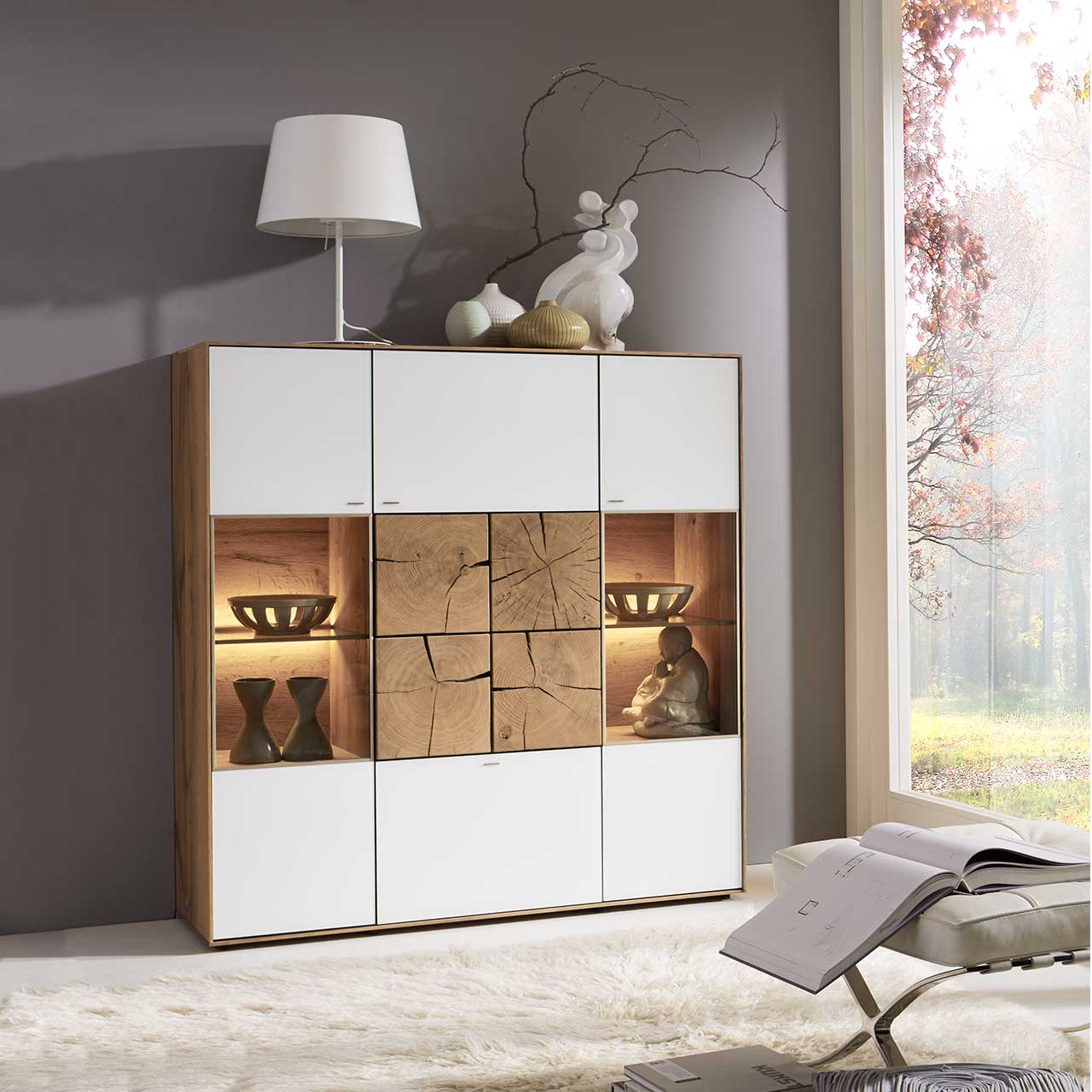 Modernes Highboard aus massiver Kerneiche Umato mit Mattglas weiß Fronten, rechts und links mit beleuchtetem Glasausschnitt, mittig mit vier dekorativen, rechteckigen Hirnholzscheiben in heller Kerneiche Umato - in einem modernen Flur stehend.