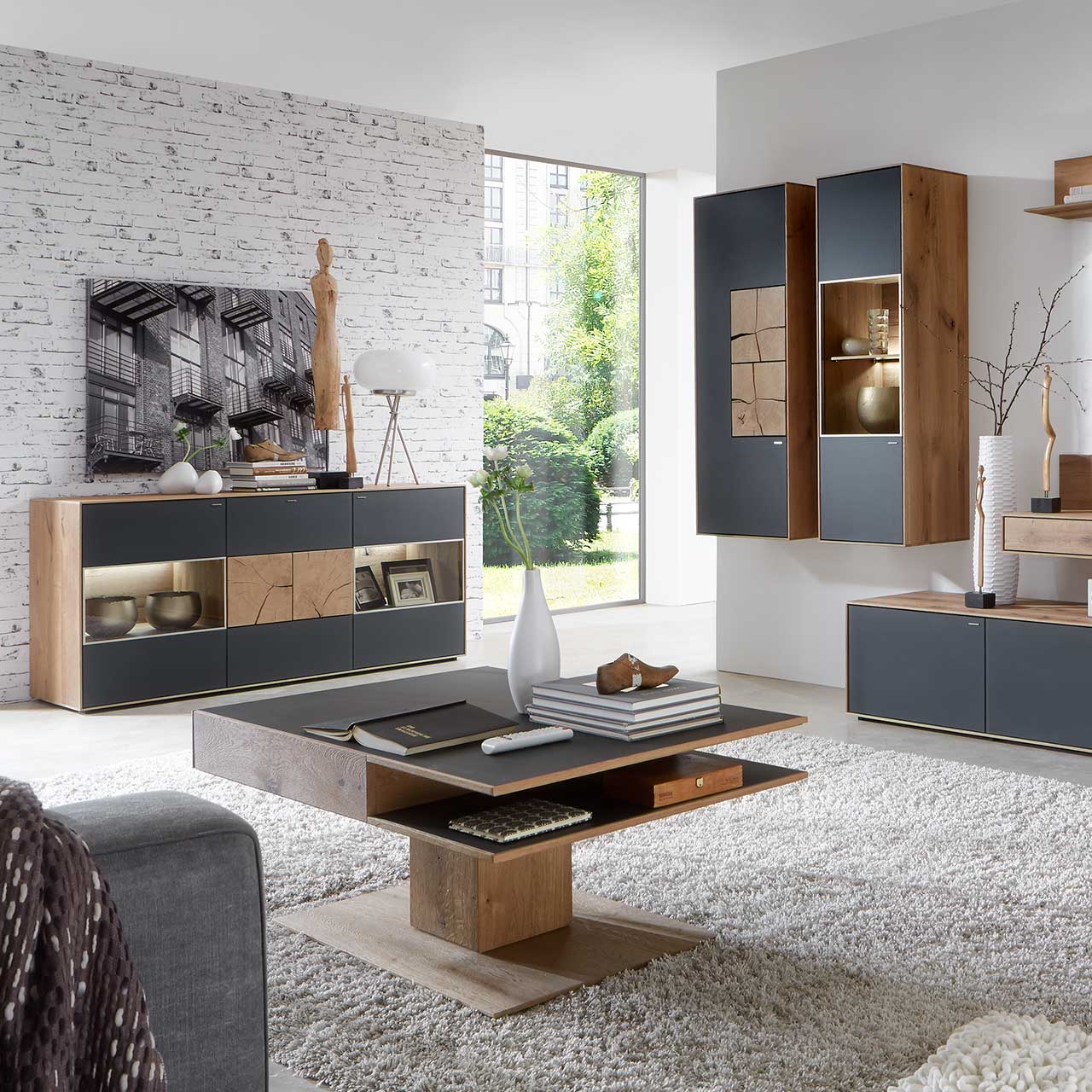 Milieubild Hartmann CAYA - Couchtisch 7170-0446 Wohnzimmer mit modernen Massivholzmöbel aus Kerneiche Umato mit Mattglas anthrazit Fronten: Hängeschrank mit Hirnholzscheiben, beleuchtete Hängevitrine, links Sideboard mit Glasfront und Hirnholzscheiben, davor rechteckiger Holz-Couchtisch mit Ablagefach.