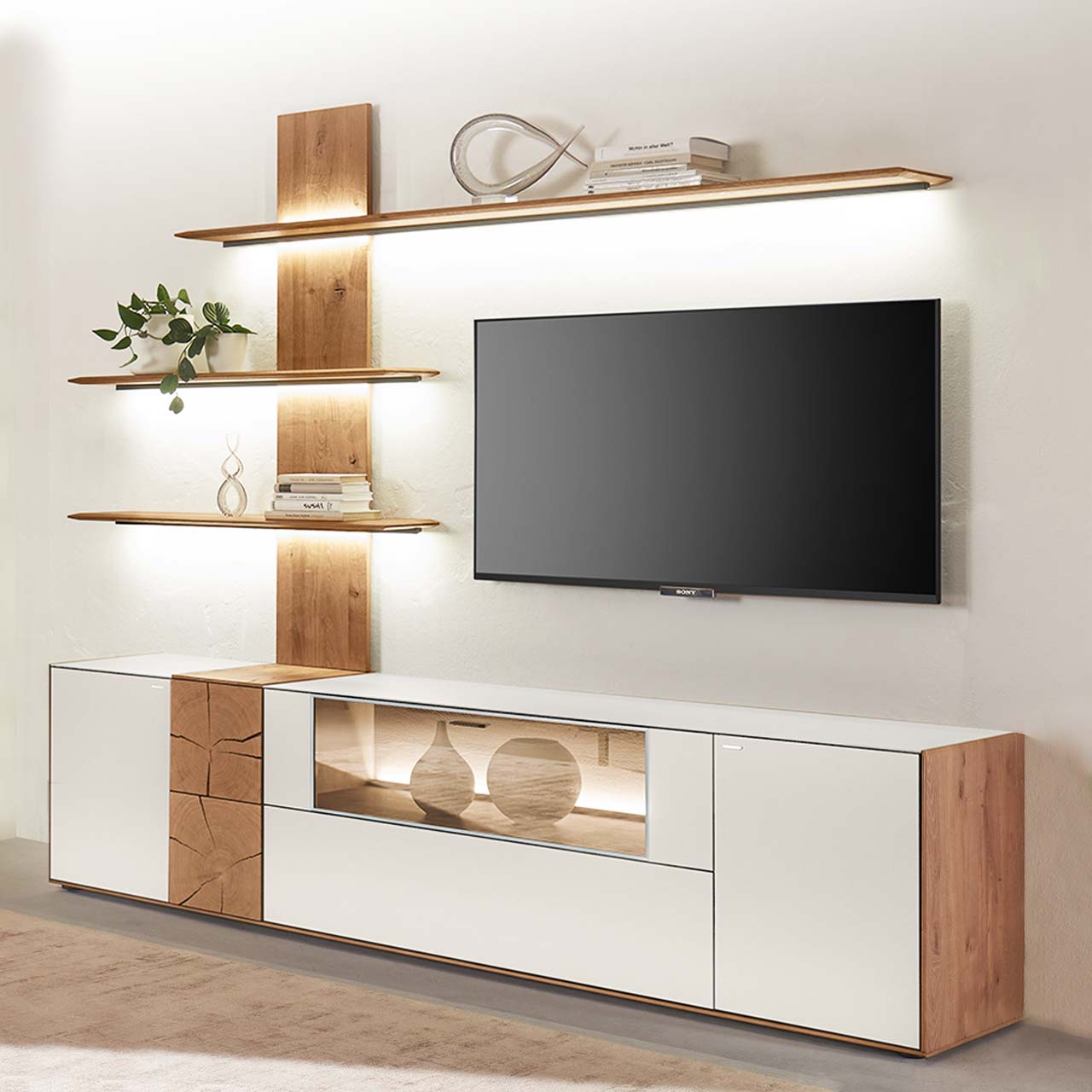 Moderne zweiteilige Massivholz Wohnkombination aus Kerneiche Umato mit Mattglas weiß Fronten und Hirnholzscheiben, mit einem langen Lowboard mit Glasausschnitt und einem Wandpaneel mit drei beleuchteten Regalböden. Dazwischen hängender Fernseher.