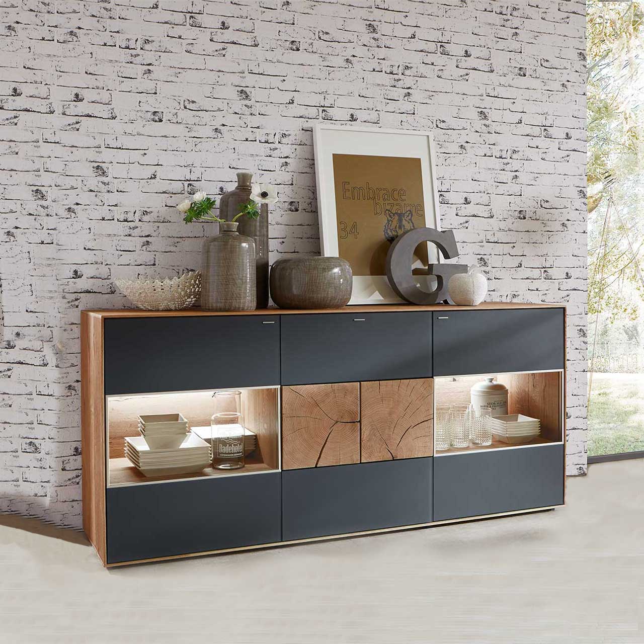 Milieubild Hartmann CAYA - Sideboard mit Glastüren 7170-4174A Modernes Sideboard aus massiver Kerneiche Umato mit anthrazitfarbenen Mattglasfronten. Rechts und links mit beleuchtetem Glasausschnitt, mittig mit zwei dekorativen, rechteckigen Hirnholzscheiben - vor hellgrauer Steinwand-Tapete.