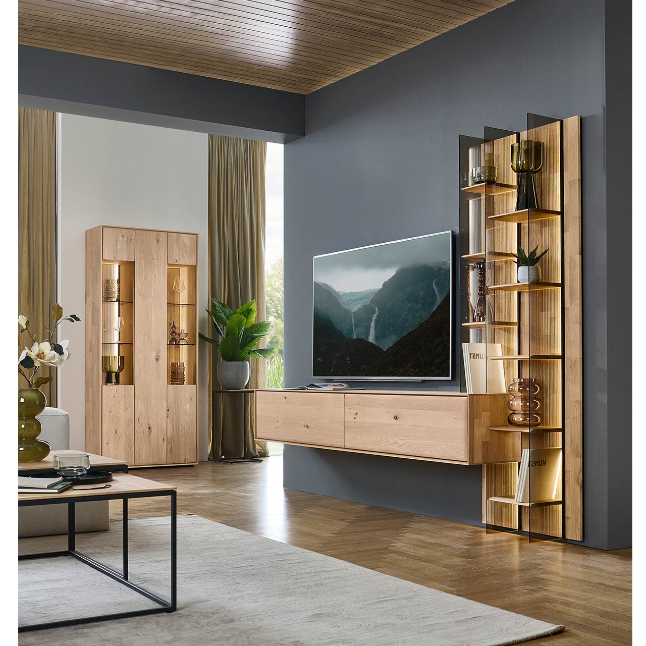 Modernes Wohnzimmer mit zweiteiliger Massivholzwohnwand mit einem hohen Wandregal mit Holzböden, beleuchteten Glasseiten und geschroppter Rückwand und einem darin integriertem Hängelowboard. Rechts an der Wand passende Standvitrine.