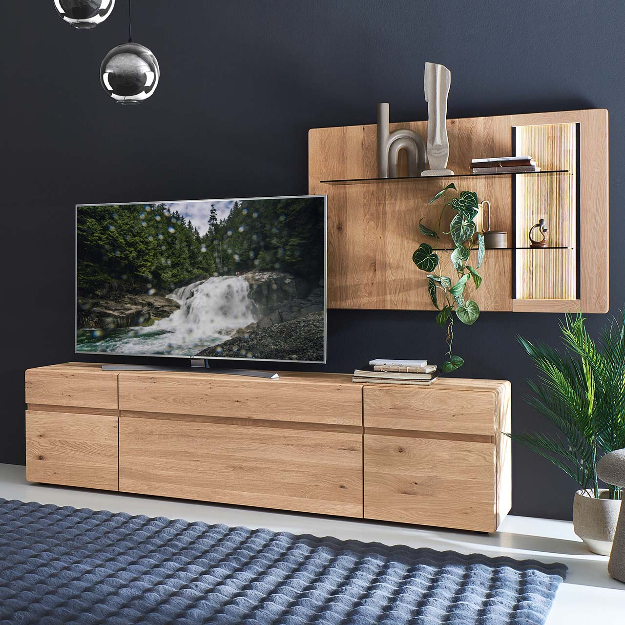 Moderne zweiteilige Wohnwand aus massiver Kerneiche Natur bestehend aus Lowboard mit Türen und Schubkasten, rechts Wandpaneel mit beleuchteten Glasböden sowie geschroppter Rückwand.