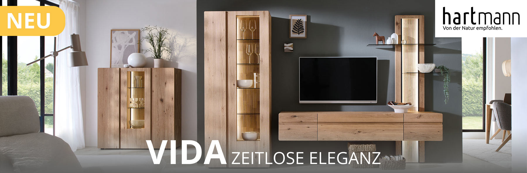 Hartmann Möbel - Möbelserie VIDA - Kerneiche Natur - Wohn-/Esszimmermöbel Hartmann VIDA - Massivholzmöbel aus Kerneiche Natur kombiniert mit Parsolglas und geschoppten Rückwänden. Ambientebild zeigt Hängelowboard, Wandpaneel mit beleuchteten Glasböden, links hohe Standvitrine, im Hintergrund passendes Highboard.
