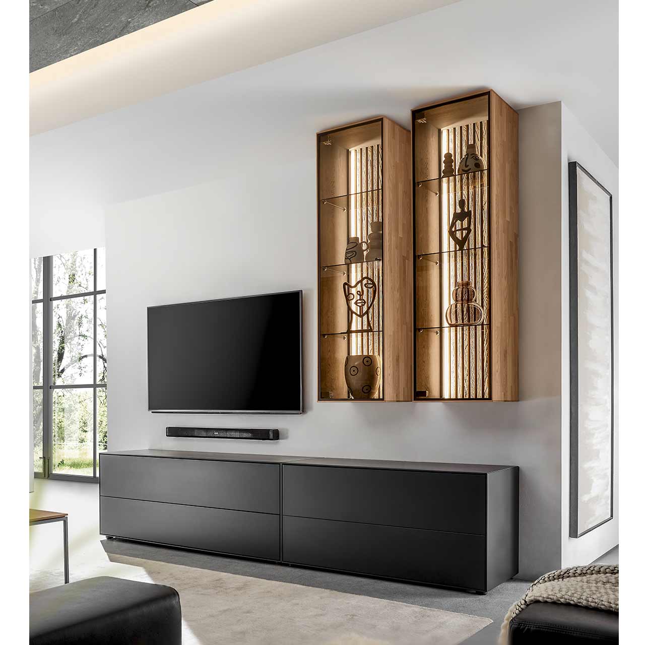 Wohnzimmer mit Fernseher und moderner Massivholz-Wohnwand mit zwei Lowboards aus Birke anthrazit und zwei Hängevitrinen aus heller Kerneiche mit beleuchteter Lamellenrückwand.