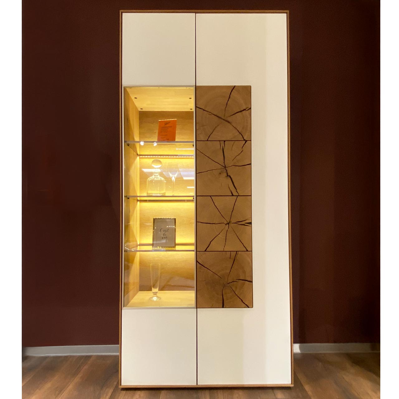 Hartmann SALE Möbel - CAYA Standvitrine 7170-0097W re. - inkl. Beleuchtung / Originalbild Ausstellung Moderne Standvitrine aus Massivholz mit weißen Mattglasfronten, rechts mit Tür und drei Hirnholzscheiben in heller Kerneiche, links mit Tür und beleuchtetem Glasausschnitt.