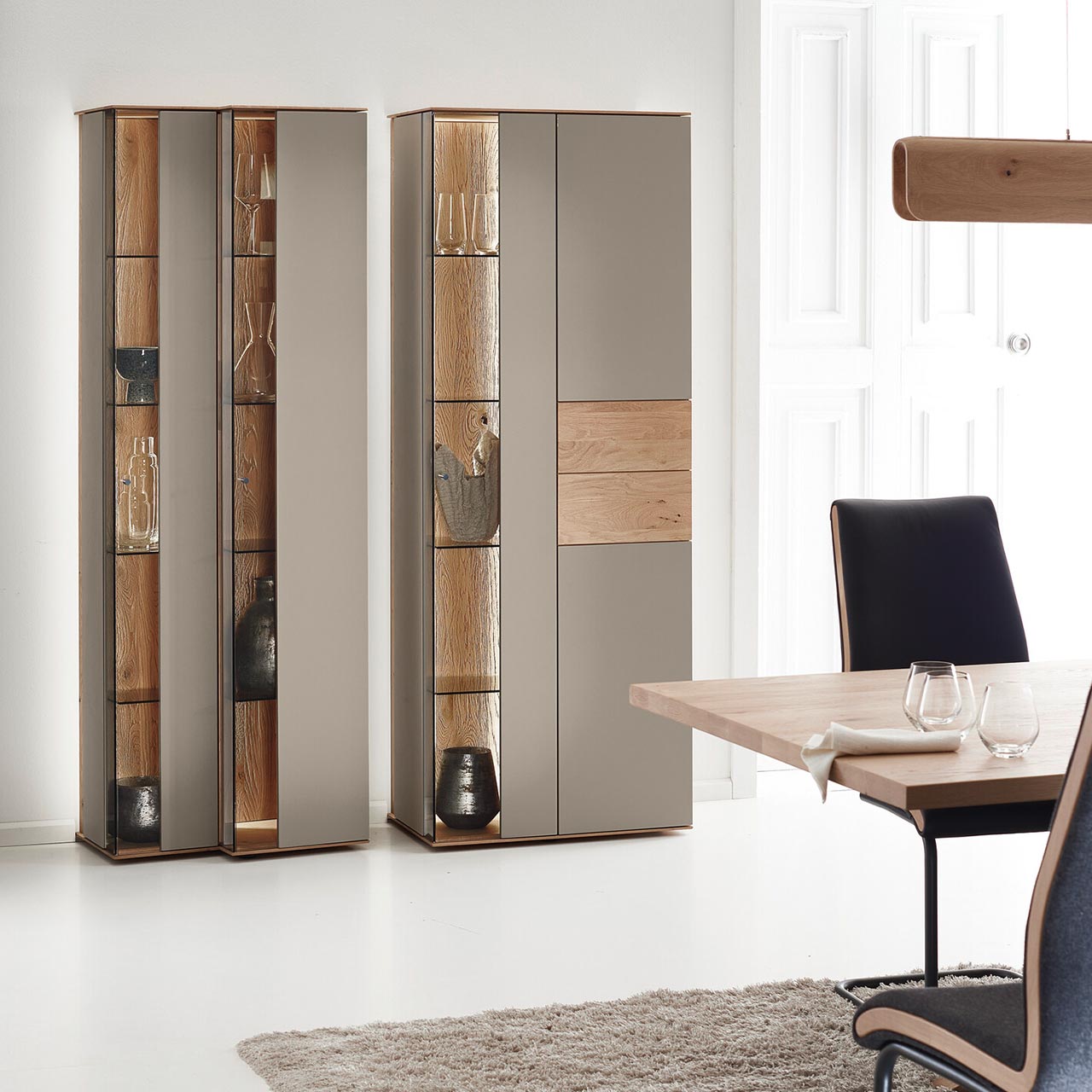 Moderne Standvitrinen aus massiver Kerneiche Natur mit Lackfronten in Sand. Mit edlen Glastüren in asymmetrischem Design und beleuchteten Riffholzrückwänden.