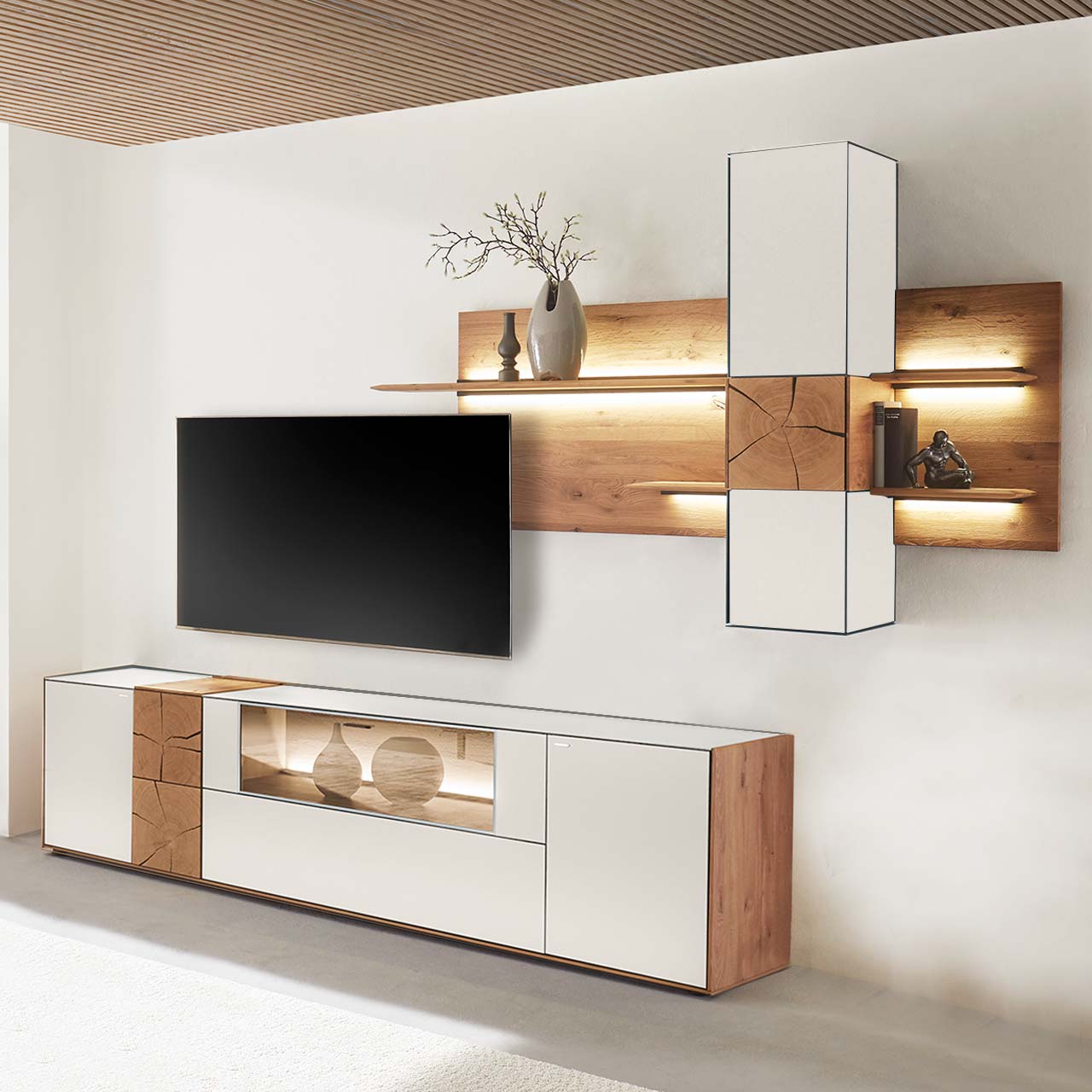 Moderne Wohnwand aus massiver Kerneiche mit Mattglas weißen Fronten. Unten ein Lowboard mit Vitrine und dekorativer Holzapplikation, oben eine Wandpaneel-Kombination mit Regalböden, Schrank und indirekter Beleuchtung, dazwischen hängender Fernseher.