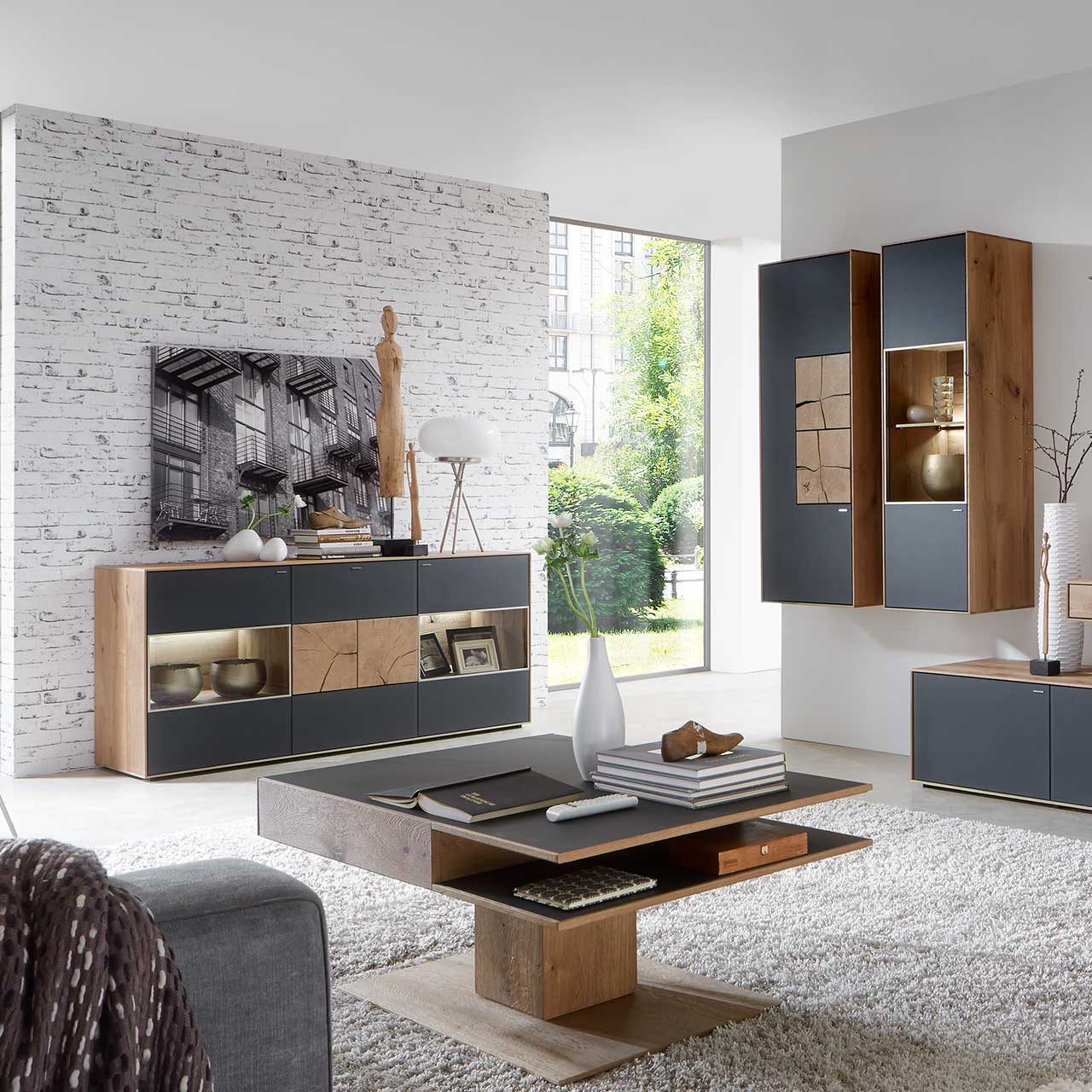 Milieubild Hartmann CAYA - Sideboard mit Glastüren 7170-4174A Wohnzimmer mit modernen Massivholzmöbel aus Kerneiche Umato mit Mattglas anthrazit Fronten: Hängeschrank mit Hirnholzscheiben, beleuchtete Hängevitrine, links modernes Sideboard mit Glasfront und Hirnholzscheiben, davor passender Couchtisch.