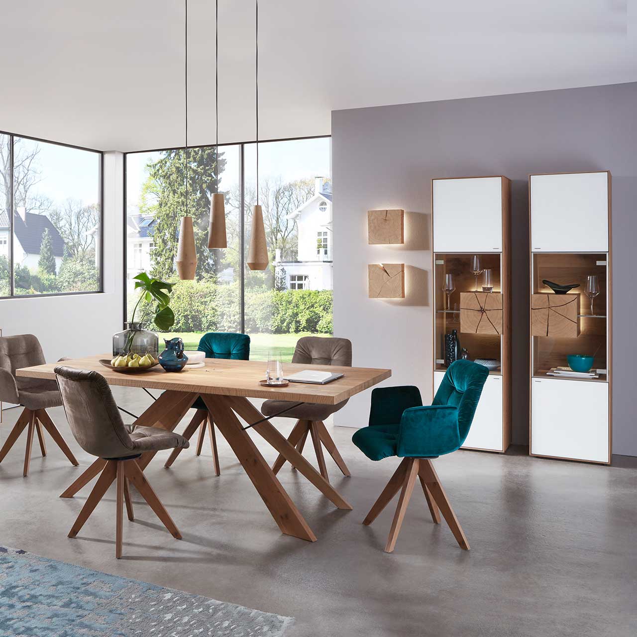 Esszimmer mit zwei modernen Standvitrinen aus massiver Kerneiche Umato mit Mattglas weiß Front, mittig mit beleuchtetem Glasausschnitt, Glasboden und Hirnholzblock. Im Vordergrund passender Holz-Esstisch und fünf Stühle mit Holzgestell.
