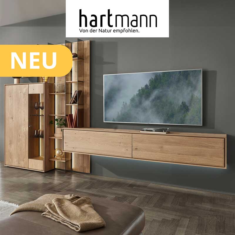 Hartmann VALA - Neue Möbelserie aus Kerneiche Natur kombiniert mit Parsolglas und geschoppten Rückwänden. Ambientebild zeigt schlichtes Lowboard, Wandpaneel mit beleuchteten Glasböden und integriertes Highboard. Weiter links Sideboard darüber Regal.