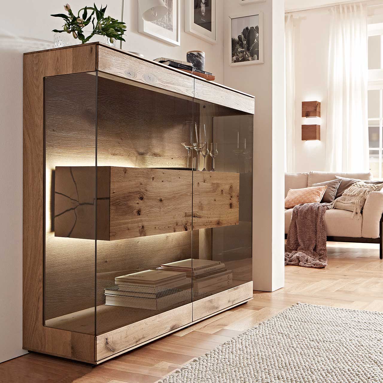 Edles Highboard aus massiver Kerneiche Umato mit großen über Ecke verlaufenden Glastüren in Parsolbronze. Innenliegend ein großer, beleuchteter Hirnholzblock mit Doppelschubkasten - in einem modernem Flur stehend.