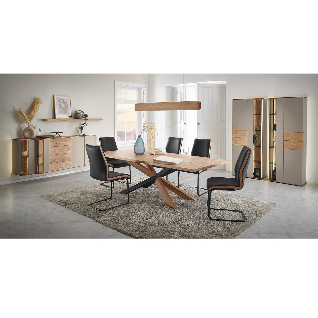 Hartmann Esszimmermöbel SVEA 8660 - aus massiver Kerneiche Natur, Lack Sand, Riffholz - Milieubild Moderne Esszimmermöbel aus massivem Holz mit Lackfronten in Sand, Parsolglas Bronze und Riffholz: Rechteckiger Holztisch mit Sterngestell, fünf Freischwinger mit dunklem Lederbezug, asymmetrisches Hängesideboard und zwei Standvitrinen in Holz/Lack.