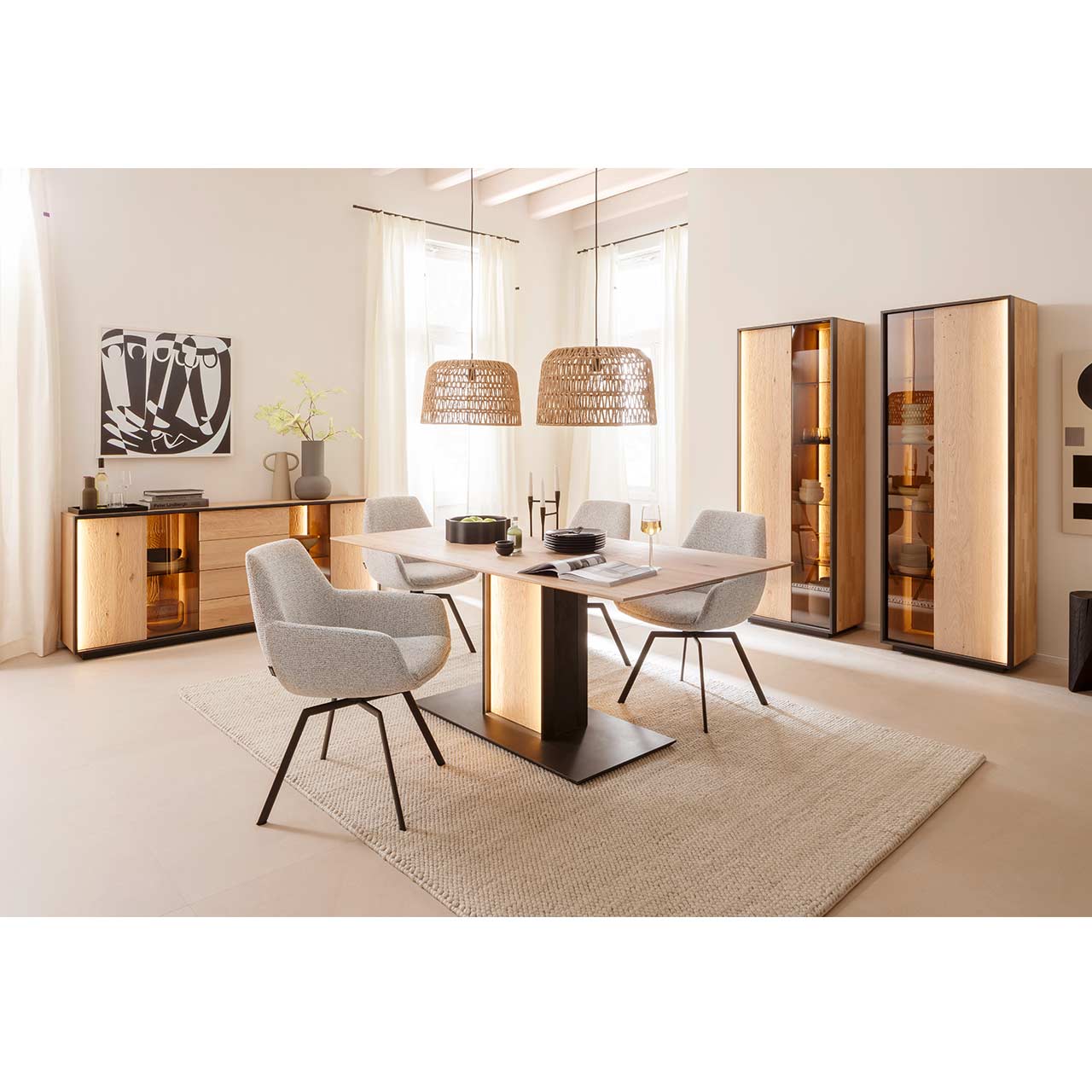 Modern eingerichtetes Esszimmer mit Massivholzmöbeln aus Kerneiche Natur mit einem rechteckigen Holztisch und vier hellen Stühlen. Links an der Wand ein Sideboard mit Glasausschnitt, rechts zwei Standvitrinen jeweils mit einer Holz-/Glastür.