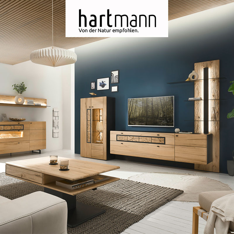 Hartmann KNUD - Massivholzmöbel aus heller Kernesche kombiniert mit anthrazit und dekorativen, eckigen Hirnholzscheiben. Ambientebild zeigt Hängelowboard mit Hirnholz an beleuchtetem Wandpaneel mit Glasböden. Links ein Highboard mit Glastür und Hirnholz.