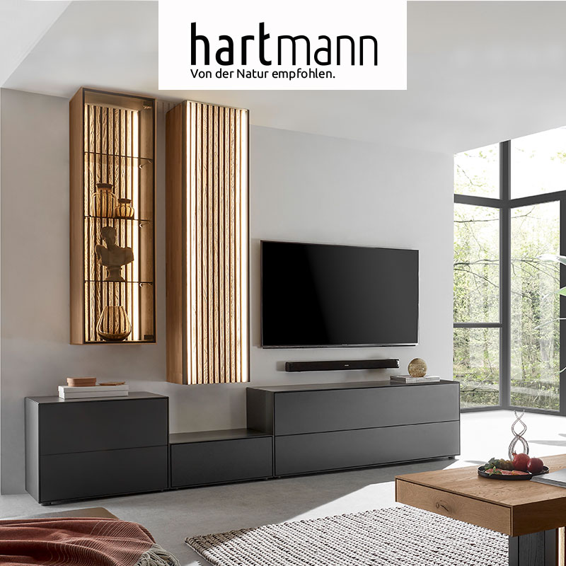 Hartmann Möbel - Möbelserie SELINO Hartmann SELINO - Massivholzmöbel aus Birke anthrazit kombiniert mit Kerneiche Natur und hellen Holzlamellen. Ambientebild zeigt drei unterschiedlich hohe, dunkle Lowboards, darüber eine beleuchtete Hängevitrine und ein Hängeschrank mit Lamellenfront.