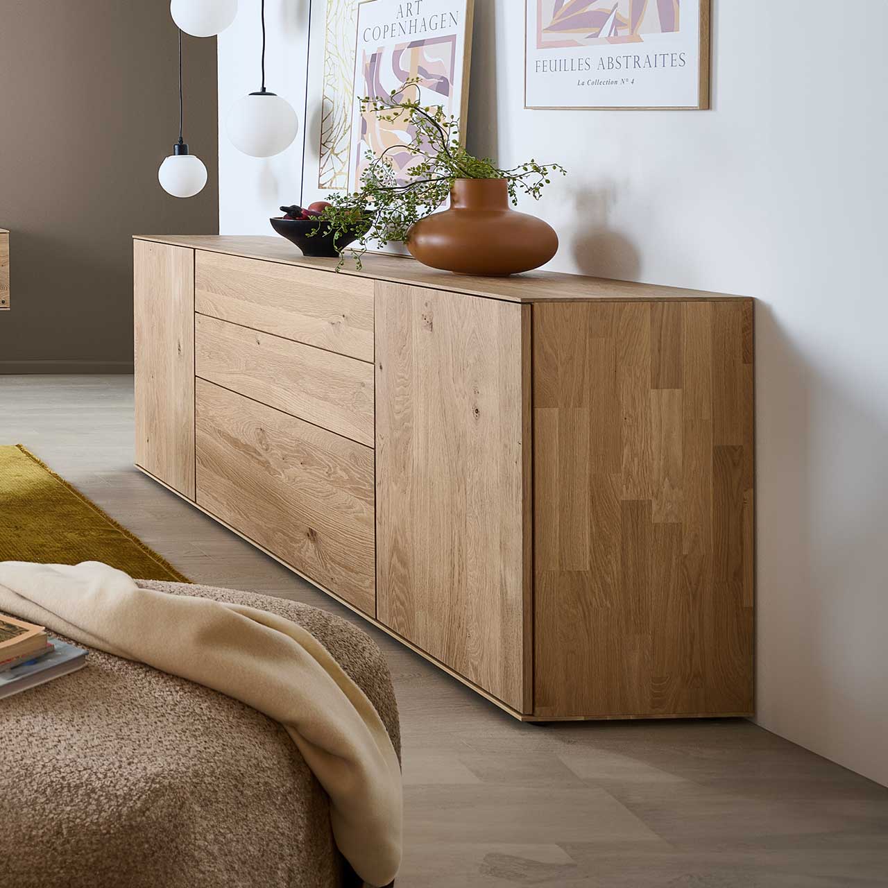 Milieubild eines seitlich platzierten Sideboards aus massiver Kerneiche Natur mit zwei Türen, Klappe und Schubkasten – vor hellgrauer Wand mit Wandbildern