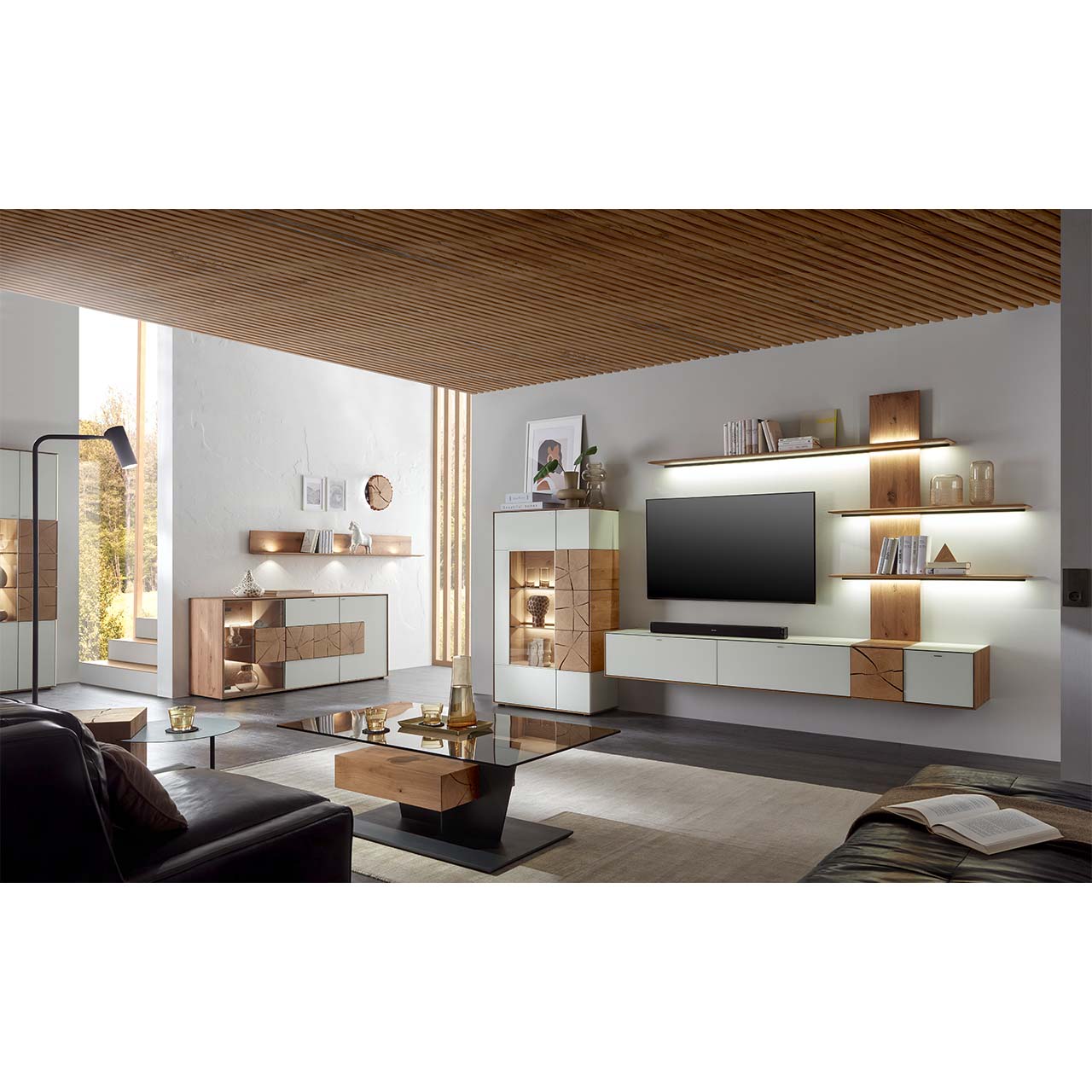 Wohnzimmer mit TV und moderner Wohnwand aus massiver Kerneiche und Mattglas weiß Fronten, mit schmalem Hängelowboard, beleuchtetem Wandpaneel und Standvitrine. Rechts ein Highboard darüber Regal. Im Vordergrund ein Couchtisch mit Glasplatte.